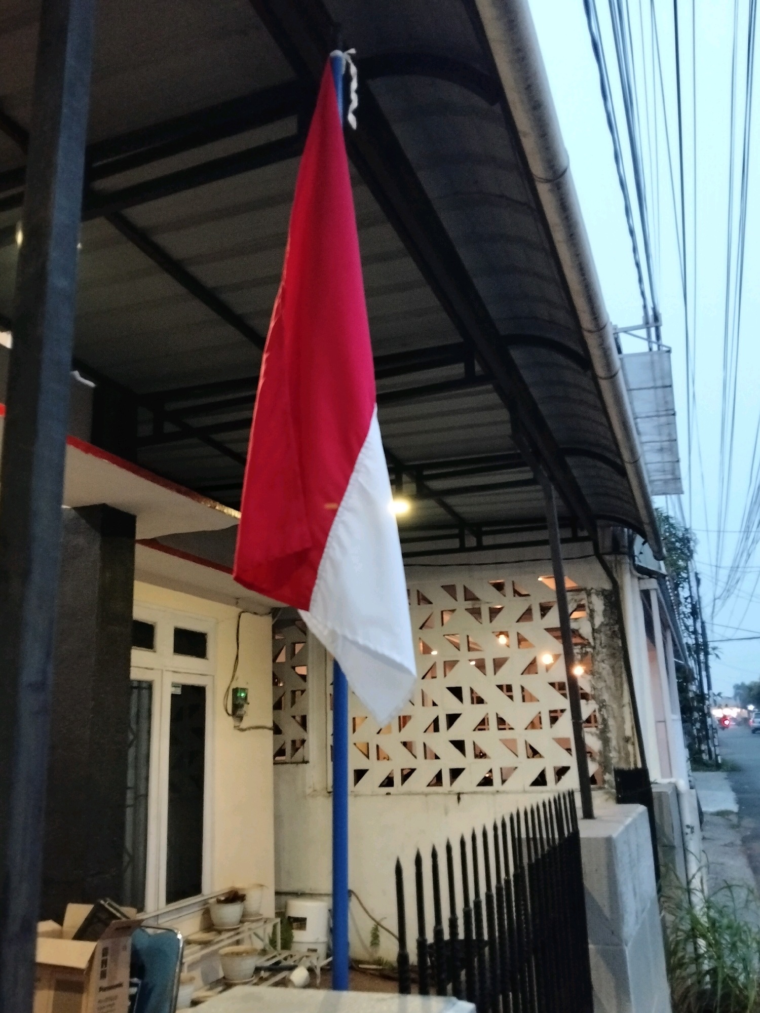 The image shows an Indonesian flag flying from a blue pole. The flag is red and white, with the red portion on top and the white portion on the bottom. The flag is attached to the pole with a white rope. The pole is located on the porch of a building. The building has a white facade and a black accent wall. There is a window on the building, and a fence in front of the porch. In the background, there are power lines and a street with cars. The scene appears to be in a residential or commercial area. The flag is likely being flown in observance of a national holiday or event. The image is taken at dusk or early evening, as the sky is a pale blue and the lights in the building are on. There are no people visible in the image.