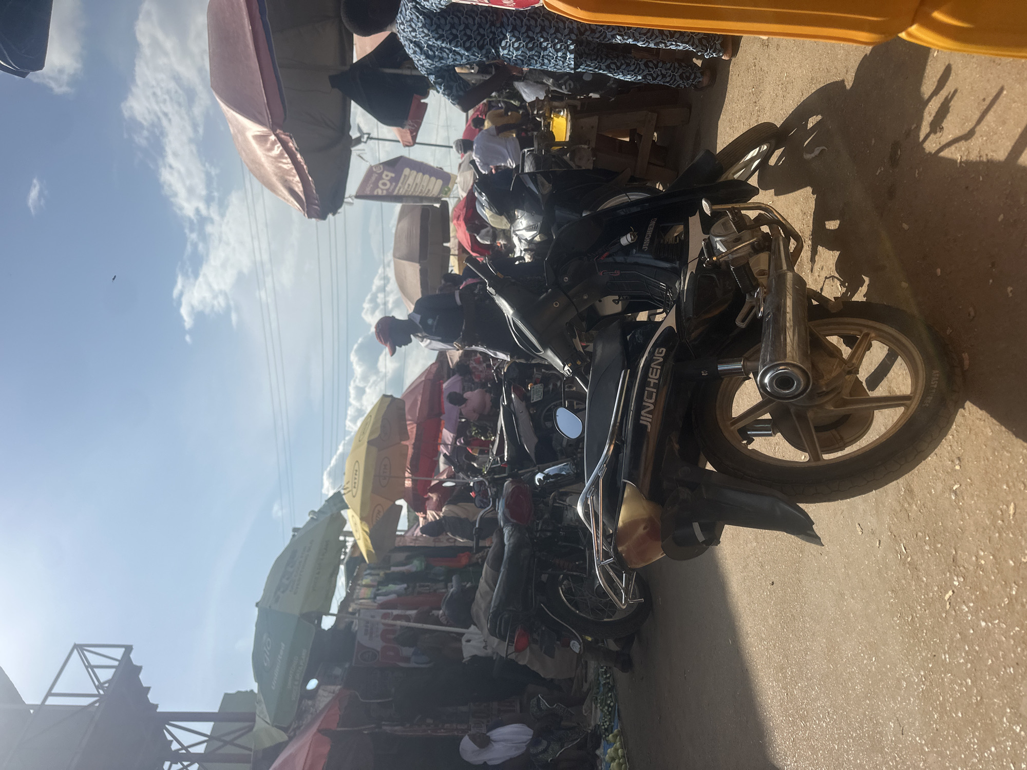 This image depicts a bustling street scene in Akwanga, Nigeria, likely a marketplace, under a bright, partly cloudy sky. In the foreground, a black motorcycle with a "JINCHENG" logo on its side is parked. The motorcycle has a visible exhaust pipe and a rear fender.
The background is filled with activity. Several other motorcycles are visible, some parked and others potentially in use. Various stalls and shops are set up, indicated by awnings and displays of goods, likely textiles or clothing, hanging behind them. Colorful umbrellas, including yellow and red ones, provide shade over some of the stalls.
People are present throughout the scene, engaged in various activities. Some individuals are seen standing, while others appear to be sitting or tending to their stalls. A man wearing a red cap and a dark shirt is standing near a motorcycle. Another person in a blue and black patterned outfit is standing in the right side of the image. A woman in a white head covering is seated on the left. The ground appears to be a dirt or unpaved surface, common in many Nigerian marketplaces. The overall impression is one of commerce and daily life in a vibrant urban setting.