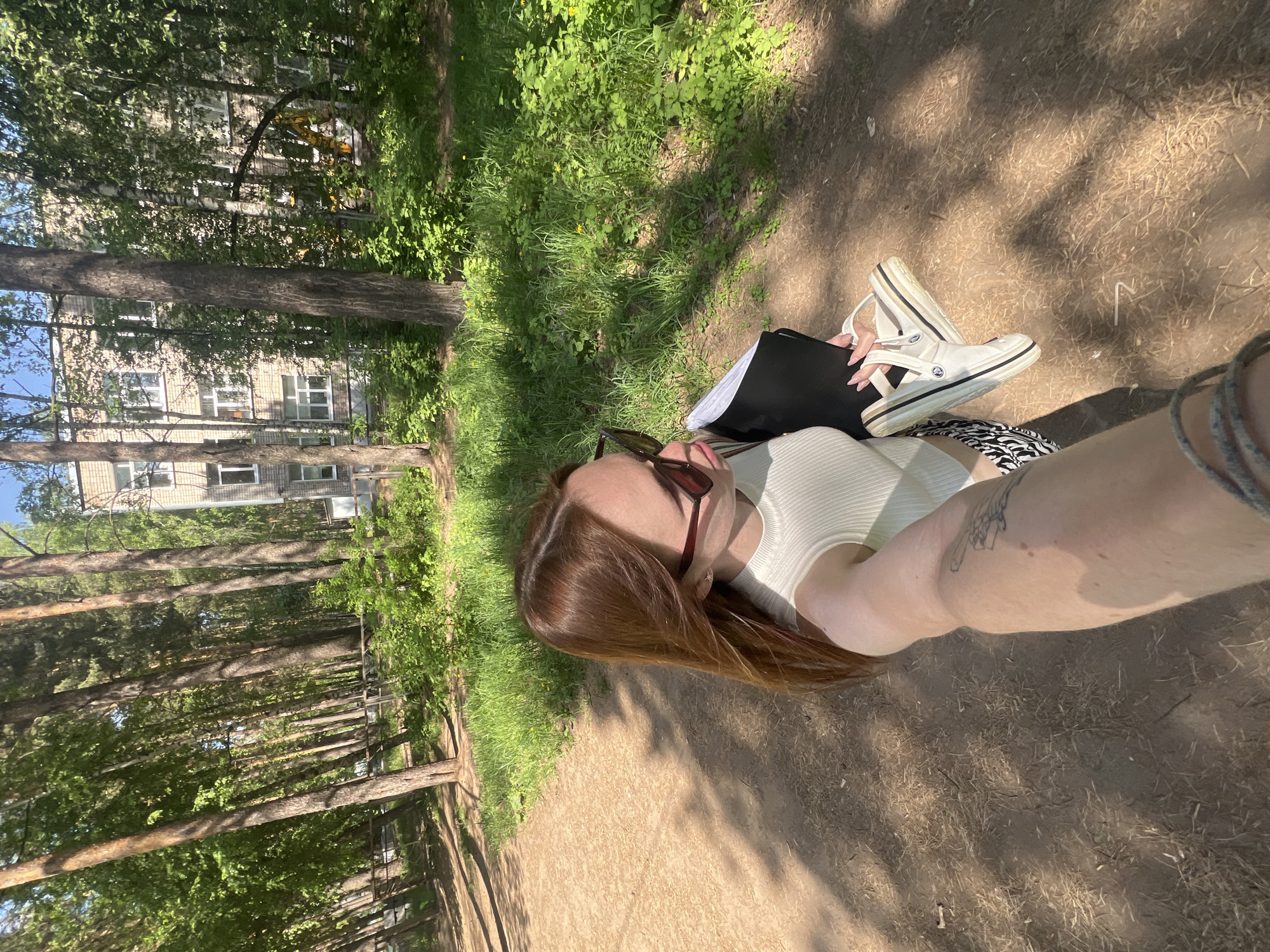 A person is sitting on a dirt path in a park-like setting. A building with multiple windows is visible through the trees in the background. The person is wearing a white tank top and patterned shorts. They are holding a pair of white shoes with black and tan accents. They also have a black portfolio or notebook. The person is wearing sunglasses and has long, red hair. There is a tattoo on their arm. The location is Chaikovsky, Russia.