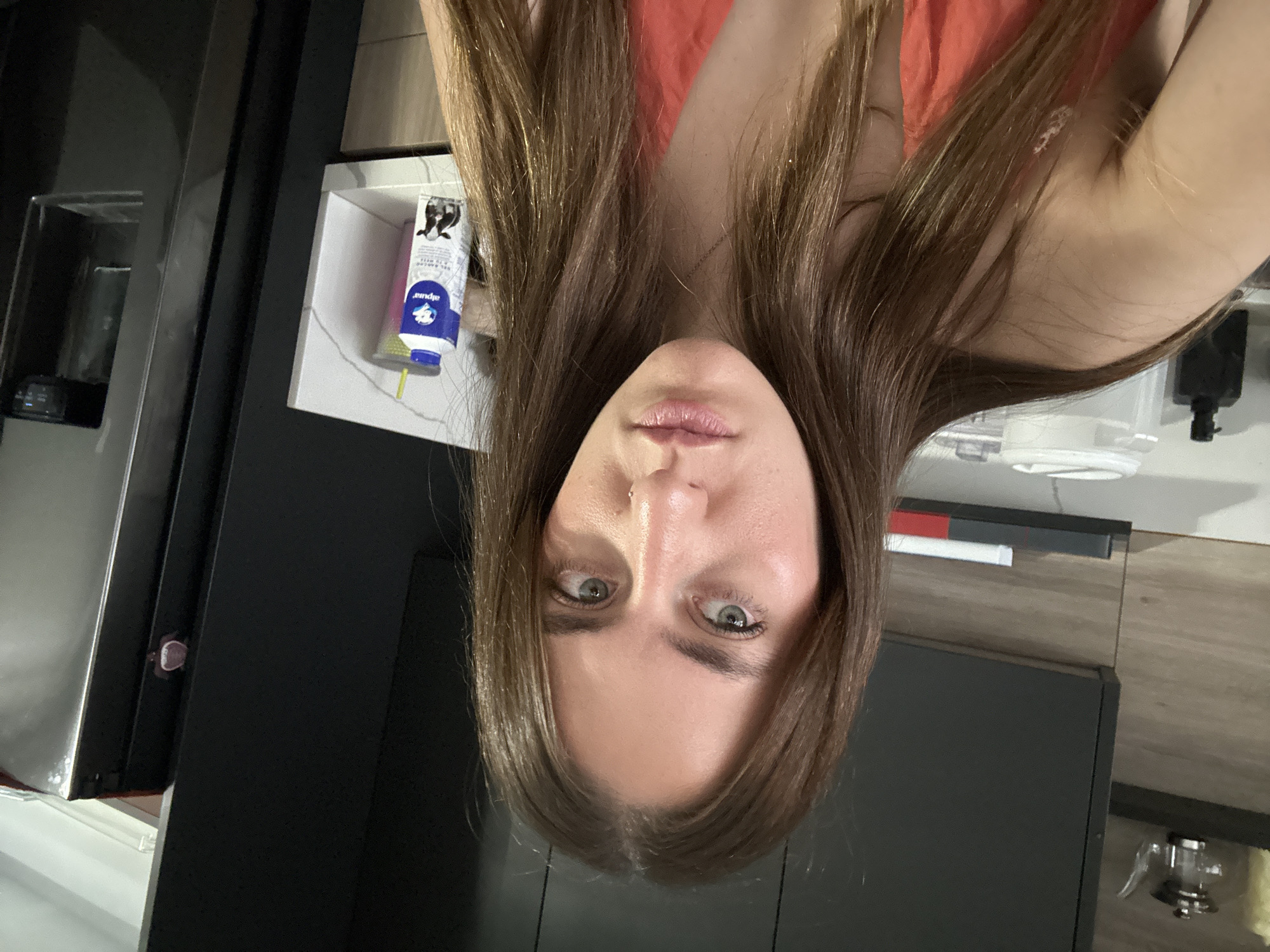 A woman with long brown hair, wearing a reddish-orange top, is positioned upside down in the center of the frame, looking directly at the camera. Her eyes are light-colored and her skin appears fair. A small nose piercing is visible.
The background consists of kitchen elements. A refrigerator is seen to the left, along with a drawer containing a small container of Alpro plant-based yogurt with a straw. Adjacent to the drawer is a countertop. Cabinetry is seen behind the woman. On the right of the countertop there is a small metal object.
The location appears to be indoors, presumably a kitchen setting. There's no explicit evidence pointing to Naucalpan de Juárez, Mexico, in the image.