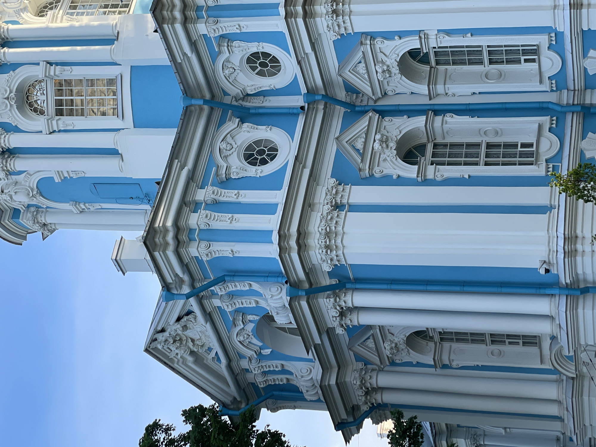The image displays a detailed, angled view of a large, ornate building, identified as being in Unknown, Unknown. The structure is characterized by its bright sky-blue walls extensively contrasted with crisp white architectural elements. These white components include numerous columns, pilasters, window frames, cornices, and intricate sculptural ornamentation.
The facade features multiple window types: tall, rectangular windows with multi-pane glazing, arched tops, and elaborate white surrounds; and circular oculus windows, each detailed with a cross-pattern mullion and framed by decorative white plasterwork that often incorporates cherubic faces (putti) at the apex. Several prominent white columns and pilasters, appearing to be in the Corinthian or Composite order, are distributed across the building's exterior, supporting elaborate cornices and architraves.
Sections of the roofline display a white pediment with complex sculptural relief. Blue downspouts, matching the wall color, are visible running vertically along the building's exterior in several locations. A small, white security camera is mounted on a blue wall segment in the lower right portion of the image. Green foliage from trees is present on the left side and bottom left, partially obscuring parts of the structure. The sky above is clear and light blue, indicating daylight conditions.