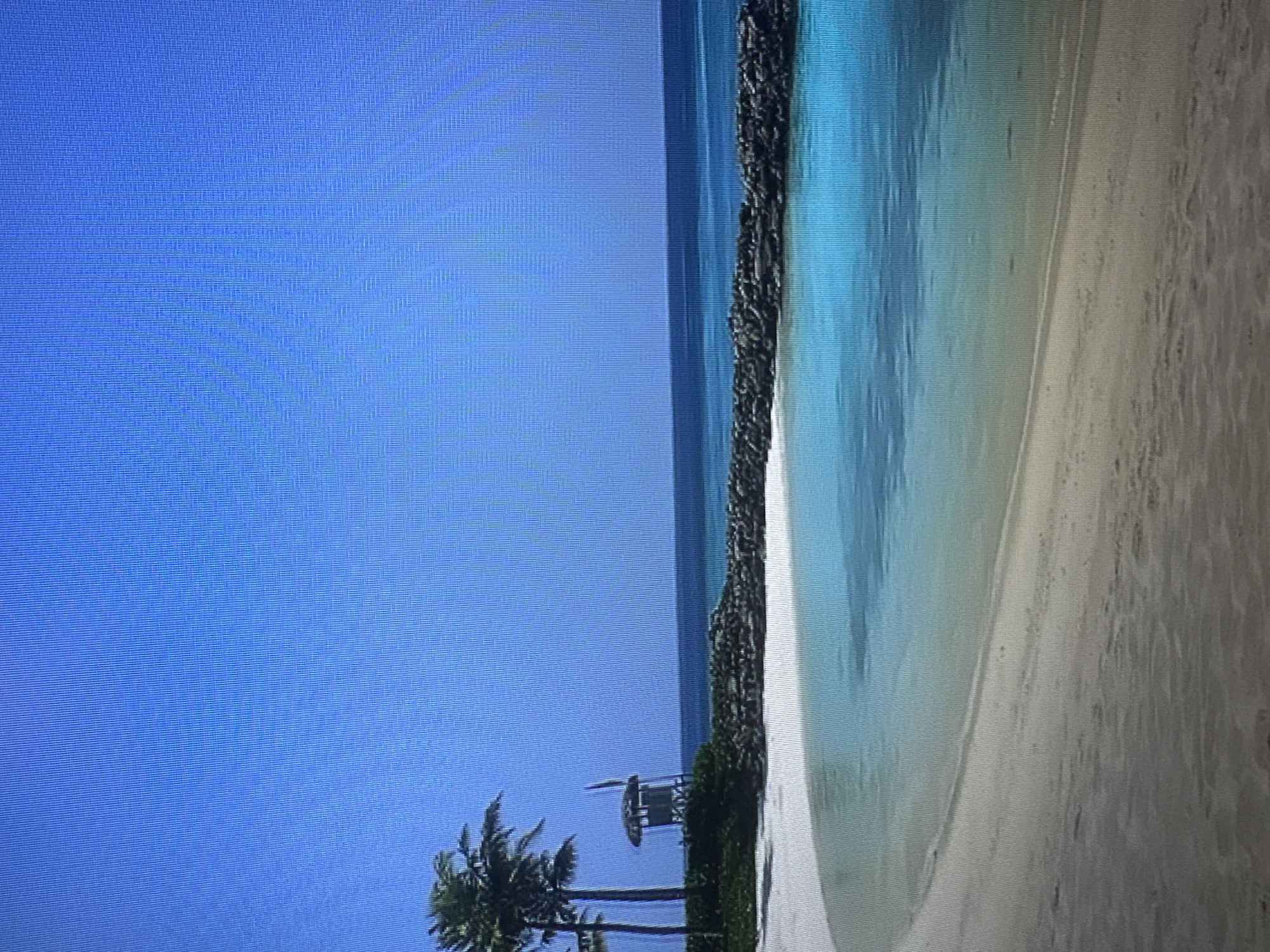 The image depicts a serene tropical beach scene under a clear blue sky. In the foreground, a gently curving white sand beach meets the shallow, clear turquoise water. The water deepens to a darker blue on the horizon. A breakwater made of dark rocks extends into the water, separating the calm lagoon from the open sea. To the left of the frame, two palm trees stand tall, and behind them is a small, rustic wooden structure, possibly a lifeguard tower or a small hut, with a thatched roof. The environment appears tranquil and undisturbed, indicative of a secluded or resort-like beach setting. This image does not depict any part of Kyiv, Ukraine.