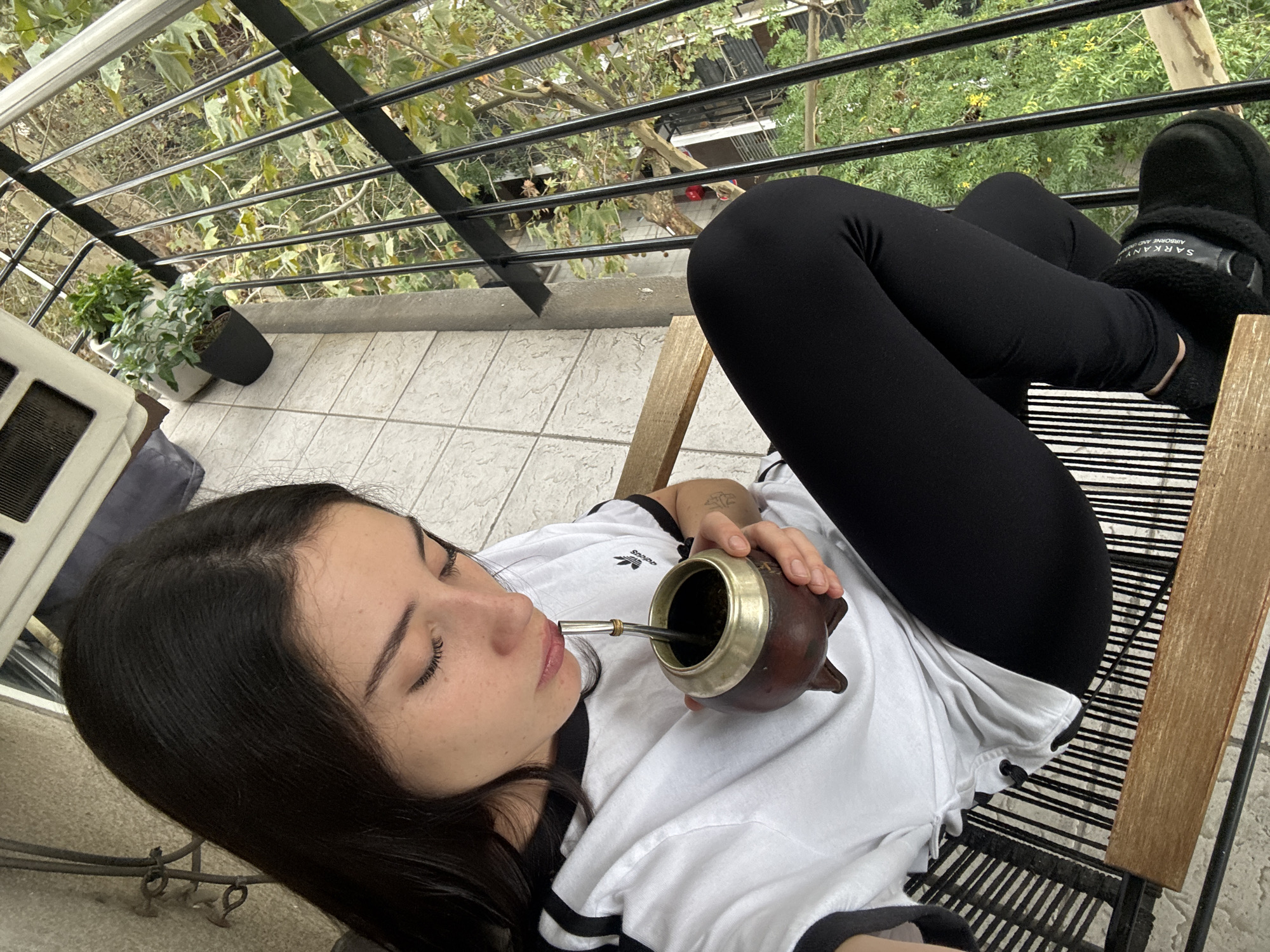 A young woman is captured relaxing outdoors on a balcony in Buenos Aires, Argentina, enjoying a moment of tranquility while drinking mate. She is lying on a slatted chair, dressed in a white t-shirt with black stripes on the sleeves and an Adidas logo, paired with black leggings. Her feet are clad in black furry slippers, one of which displays "SARKANY AIRBORNE AND CO." text. With her eyes gently closed, she brings a traditional brown mate gourd with a metal rim and silver bombilla to her lips.
The setting is a balcony with a light-colored tiled floor and a black metal railing. Beyond the railing, abundant green trees with autumnal leaves are visible, suggesting an urban residential environment. Potted plants are positioned near the railing on the balcony floor, adding a touch of greenery. Part of a white air conditioning unit or outdoor appliance is visible on the left side of the frame. The natural lighting suggests it is daytime, with soft, indirect light creating a calm atmosphere. The overall scene depicts a peaceful moment of daily life and relaxation.