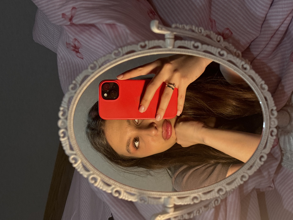 A mirror reflects a selfie taken by a person holding a red phone. The person's face, hand, and phone are visible in the mirror's reflection. The mirror's frame is ornate and white, and the background includes a striped fabric with a subtle butterfly pattern. The light is soft and highlights the person's features.
