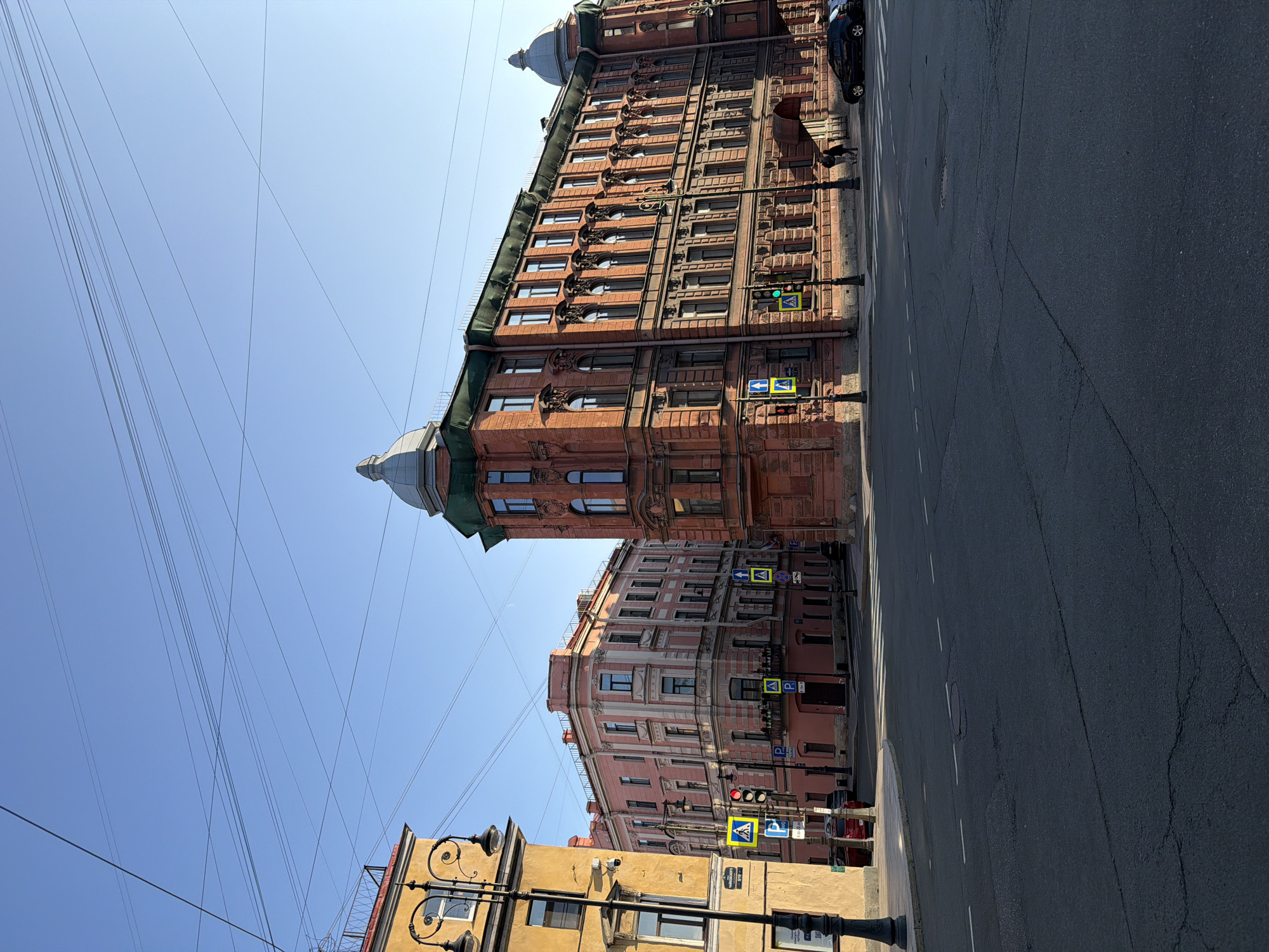 The image shows a street intersection in Saint Petersburg, Russia, on a clear day with a bright blue sky. Two large, ornate buildings dominate the scene. The building on the right is a reddish-orange brick structure with multiple floors, arched windows, and decorative architectural elements. It has a green-domed cupola at one corner and another at a further point along its facade. The building on the left is a lighter pink color with white accents, also featuring multiple floors and numerous windows. A yellow building with a decorative railing on its upper level is partially visible on the far left.
The intersection is paved with asphalt and has painted road markings, including a crosswalk. Several traffic signals and street signs are present, including pedestrian crossing signs with a yellow background and a pedestrian icon, and parking signs. A dark-colored car is parked on the right side of the street. Two figures are walking on the sidewalk in front of the reddish-orange building. A black lamppost with decorative elements stands on the left side of the intersection. Power lines are strung across the sky, crossing from one side of the frame to the other. The overall impression is of an urban environment with historic architecture.