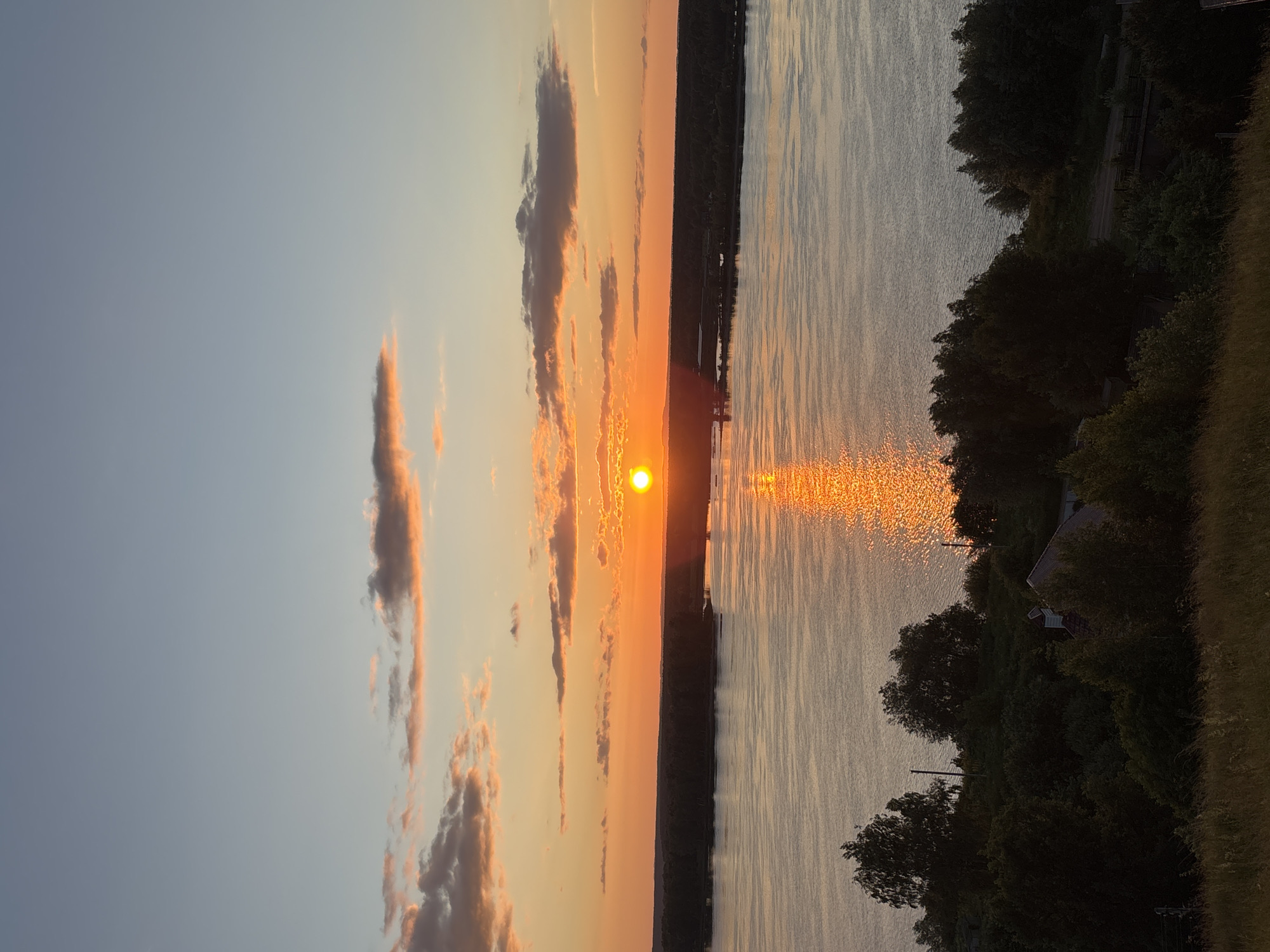 The image depicts a serene sunset over a wide river. The sun, a bright orb of orange, is low on the horizon, casting a shimmering golden path across the water. The sky is a gradient of soft blues and oranges, with a few wispy clouds tinged with pink and yellow. The far bank of the river is lined with a dense, dark green forest. In the foreground, a hilly landscape is covered with lush green trees and vegetation, with hints of a small building nestled among the foliage. The scene is one of natural beauty and tranquility. This image was captured in the city of Olkhovka, Russia.