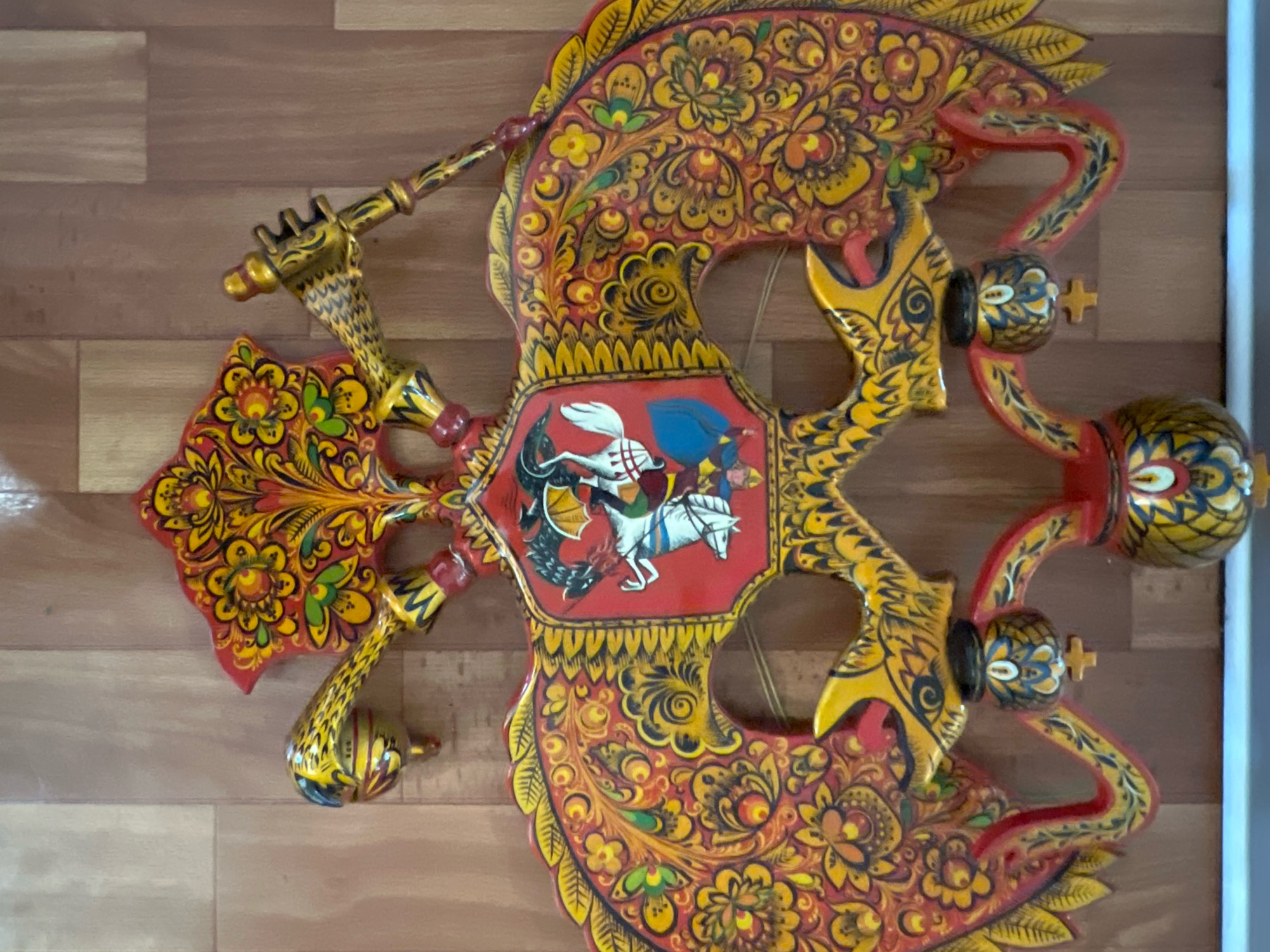 An ornate, hand-painted wooden decorative object, likely a representation of a mythical creature or a coat of arms, is displayed against a wooden paneled wall. The object is predominantly red and yellow, adorned with intricate floral patterns and detailing in black. In the central shield, a figure on horseback, possibly Saint George, is depicted spearing a dragon. The object has multiple protrusions and decorative elements, including what appear to be heads of creatures and spherical accents. This item is located in the city of Volzhsky, Russia.