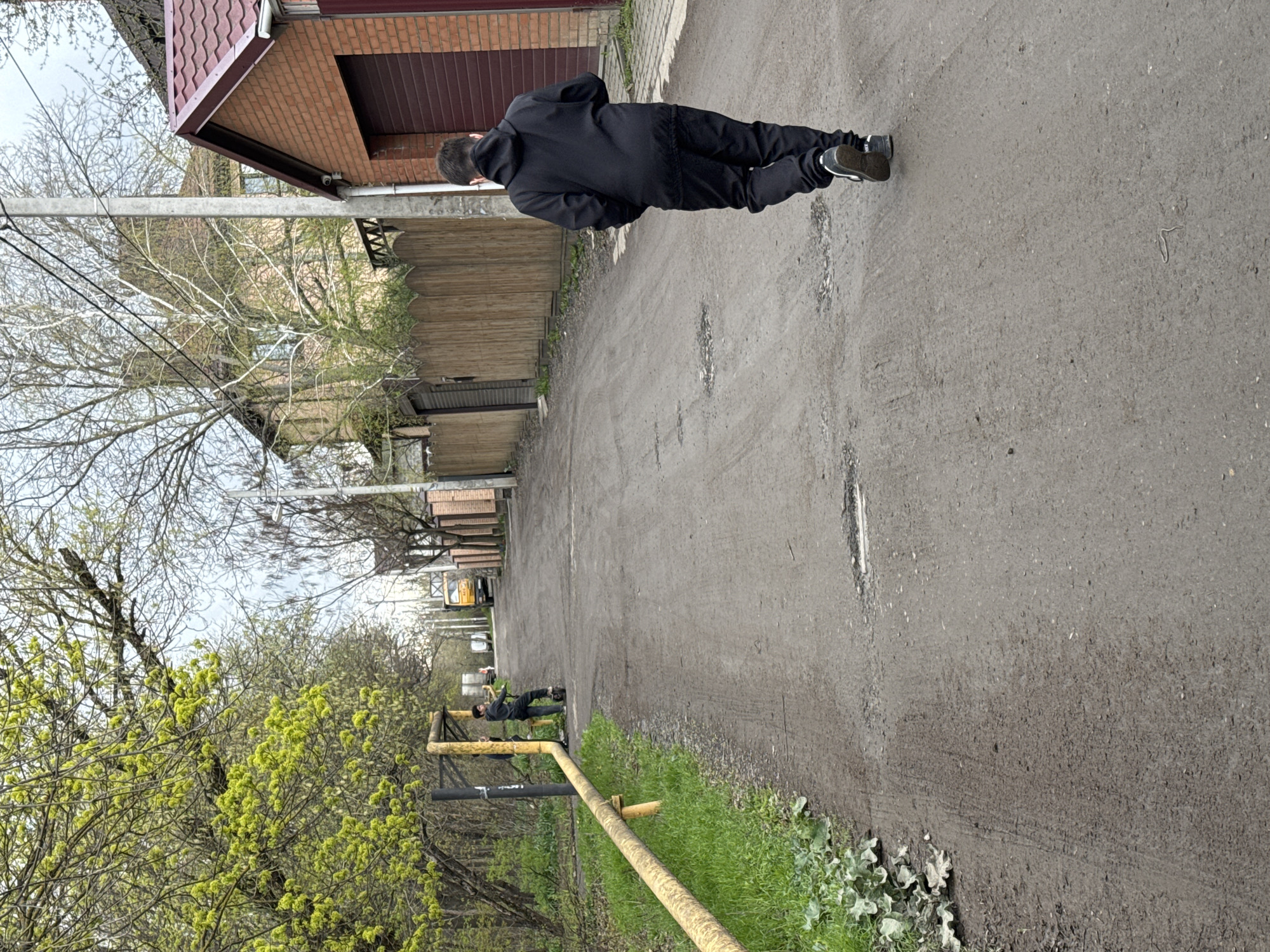 The image depicts a man in a black hooded sweatshirt and black pants walking away from the camera on a dirt road. The road is flanked by wooden fences, trees, and a brick building on one side, and greenery with a yellow barrier on the other. In the distance, another person appears to be leaning on the yellow barrier, and a yellow school bus is visible. The location appears to be Rostov-on-Don, Russia.
