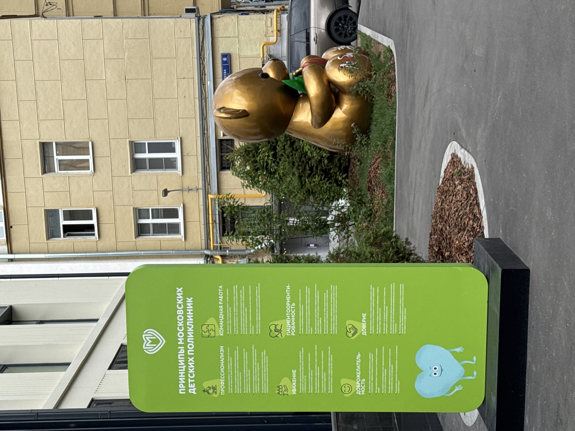 The image depicts a green sign with the title "ПРИНЦИПЫ МОСКОВСКИХ ДЕТСКИХ ПОЛИКЛИНИК" (Principles of Moscow Children's Polyclinics) and several icons with corresponding text. The sign is positioned outdoors, likely on a sidewalk or paved area, with a building in the background.
To the right of the sign, a large, golden sculpture of a teddy bear is prominently featured, seated and holding a green object. Behind the sculpture, a beige building with several windows is visible, along with some green foliage and a portion of a dark-colored SUV. A street sign with the number "6" and text in Cyrillic is also visible on the building.
The overall environment appears to be an urban setting in Moscow, Russia, characterized by the architecture and the presence of a public sculpture. The scene is captured from a slightly elevated perspective, looking down towards the sign and sculpture.