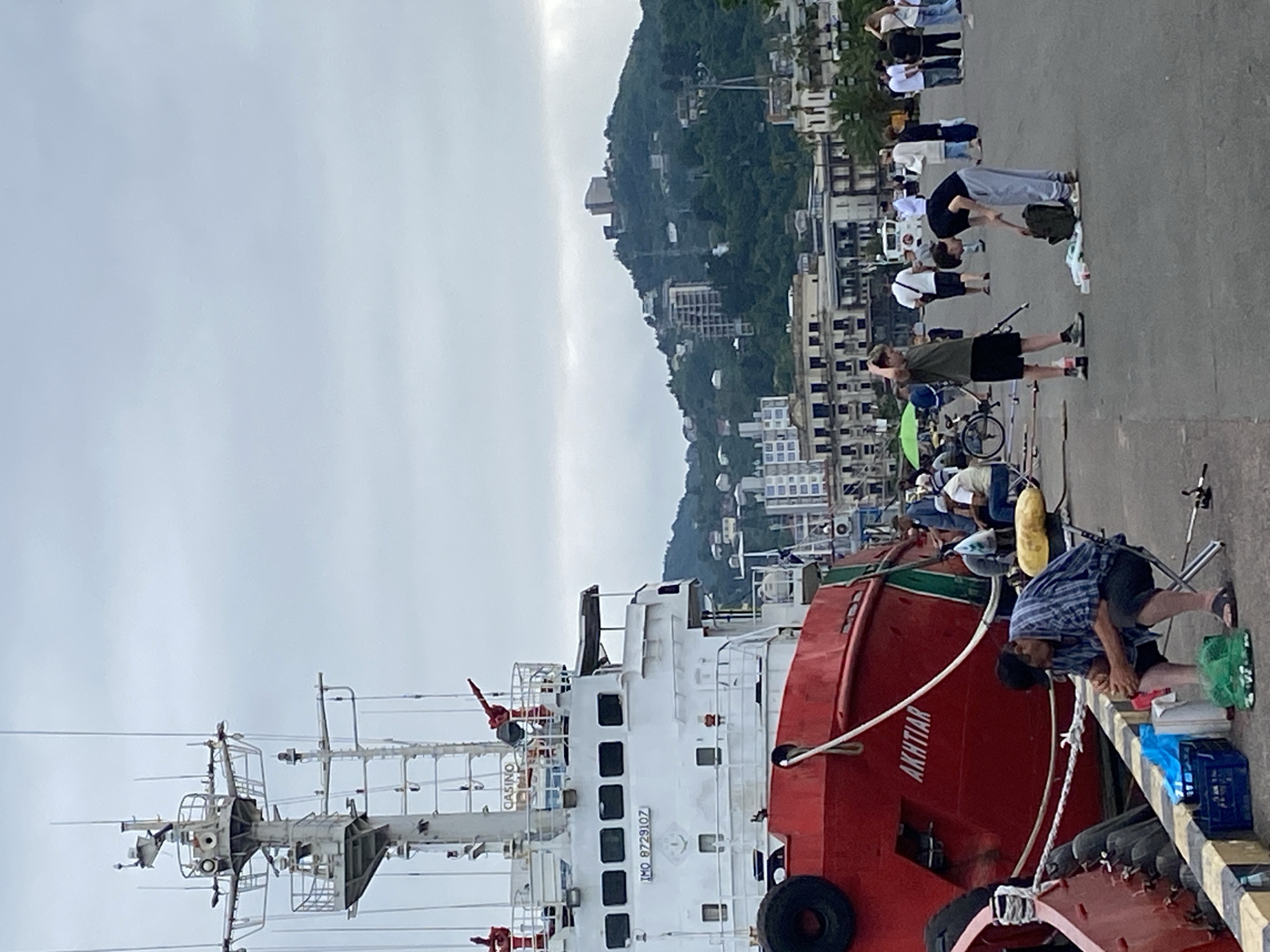 The image shows a dockside scene in Batumi, Georgia, with a large red and white ship docked. Several people are gathered on the dock, some engaged in fishing. In the background, a hillside is covered with buildings and greenery, suggesting an urban coastal environment.
The people are casually dressed, and some are actively fishing with rods. Others are standing or sitting, observing or socializing. One person is bending over a fishing tackle box. The overall atmosphere appears relaxed, possibly during a leisure activity.
The weather seems overcast, with a bright sky hinting at daylight. The prominent text visible on the ship is "AKHTIAR" and a registration number "IMO 8729107", along with "CASINO" potentially indicating the ship's purpose or a previous designation.
The scene captures a moment of everyday life by the sea, with a blend of maritime and recreational elements. The detailed infrastructure of the ship and the surrounding buildings on the hill provide context about the location.