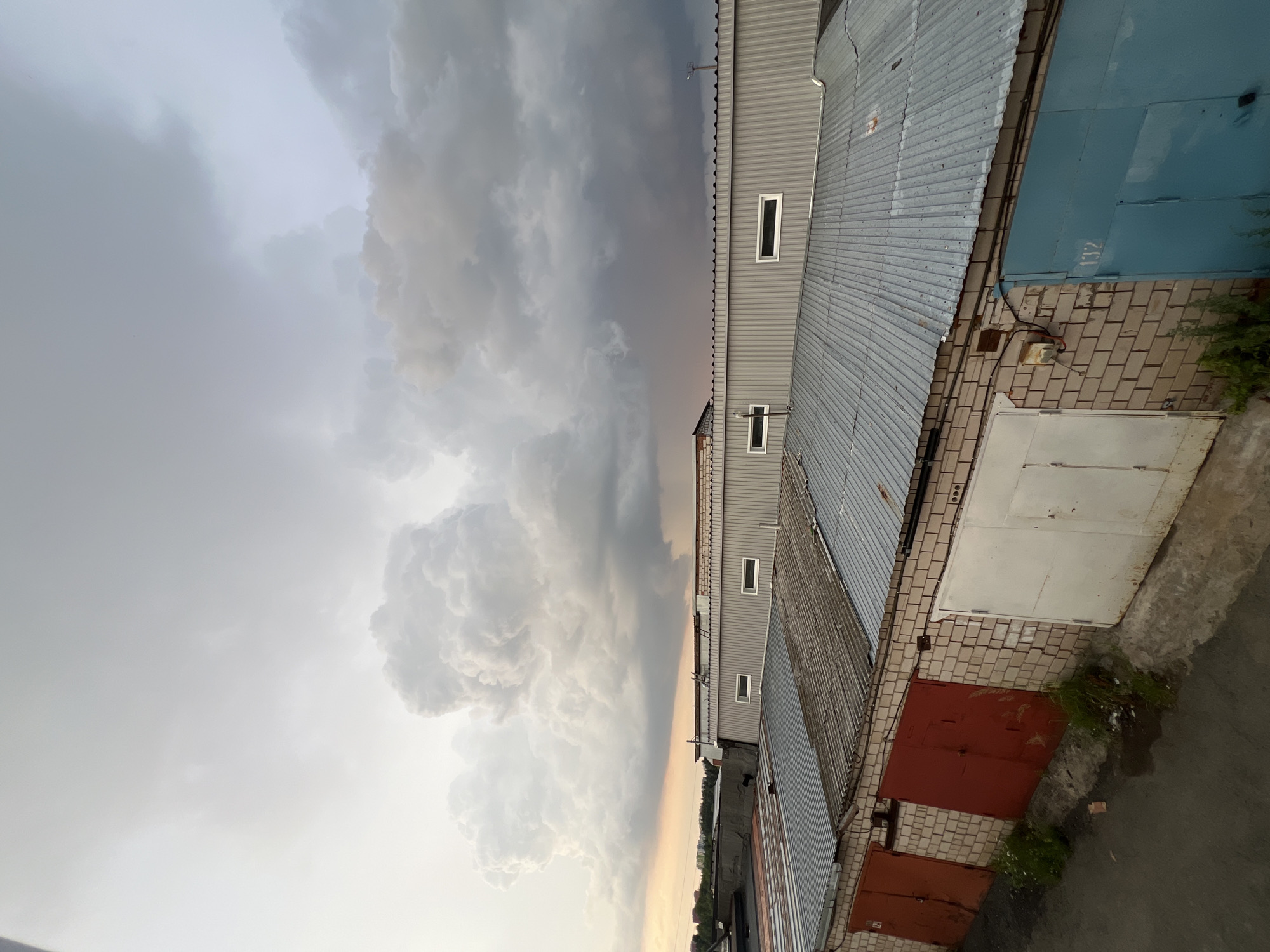 The image captures a dramatic sky filled with large, billowing cumulonimbus clouds, suggesting an impending storm or the aftermath of one. The clouds are predominantly white and gray, with some areas appearing darker, indicating heavy precipitation.
Below the imposing sky, a row of garages forms the foreground. The garages have metal roofs and brick walls. Some of the garage doors are red, one is white, and another is a striking blue. One of the blue garage doors has the number "132" stenciled on it. The garages appear to be part of a larger structure, possibly a garage complex.
There are no people visible in the image. The scene is set outdoors, likely in an urban or suburban area given the presence of garages. The lighting suggests it could be late afternoon or early evening, with the dramatic clouds casting shadows and creating a moody atmosphere. The presence of greenery, possibly weeds or small bushes, growing near the garages suggests a slightly unkempt environment. The overall mood is one of powerful natural forces at play, juxtaposed with mundane human structures.