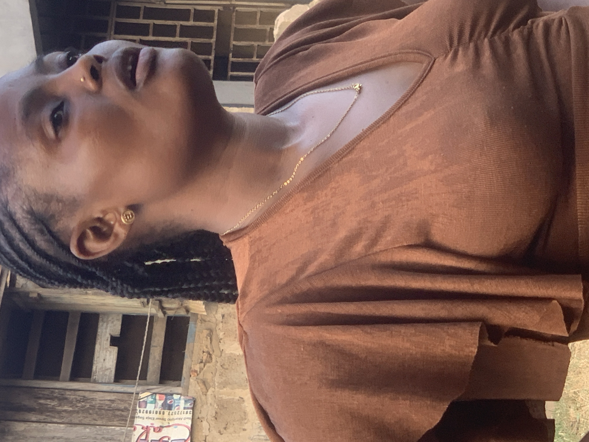A medium shot shows a Black woman in profile, looking upwards and to the right. She has dark braided hair, a gold earring in her left ear, and a gold nose stud. She is wearing a rust-colored, V-neck top with ruching at the bust and ruffled sleeves. A thin gold chain with a small pendant rests at her neck.
The background consists of a rough, stone wall with an aged, wooden structure and a barred window. To the left, a piece of paper with text and images is affixed to the wall. The lighting suggests an outdoor setting during the daytime.
This scene is located in Shagamu, Nigeria.