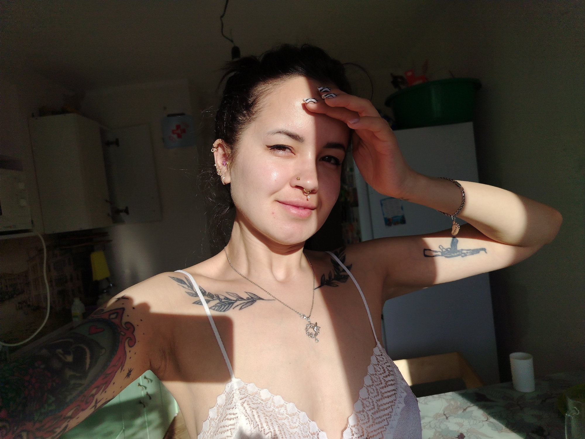 A young woman is in a sunlit room, possibly a kitchen. She has dark hair pulled back, fair skin, and multiple piercings in her ear and nose. Her fingernails are decorated with a black and white pattern. She wears a white lace camisole and a necklace. Her left arm is fully tattooed with a colorful design. A black gun tattoo is visible on her right forearm. She is holding her right hand up to her forehead, partially obscuring her face. The room includes kitchen cabinets, a refrigerator with a green plastic container on top, and a table with a floral patterned tablecloth. The location is not discernible as Noviy, Russia from visual information.