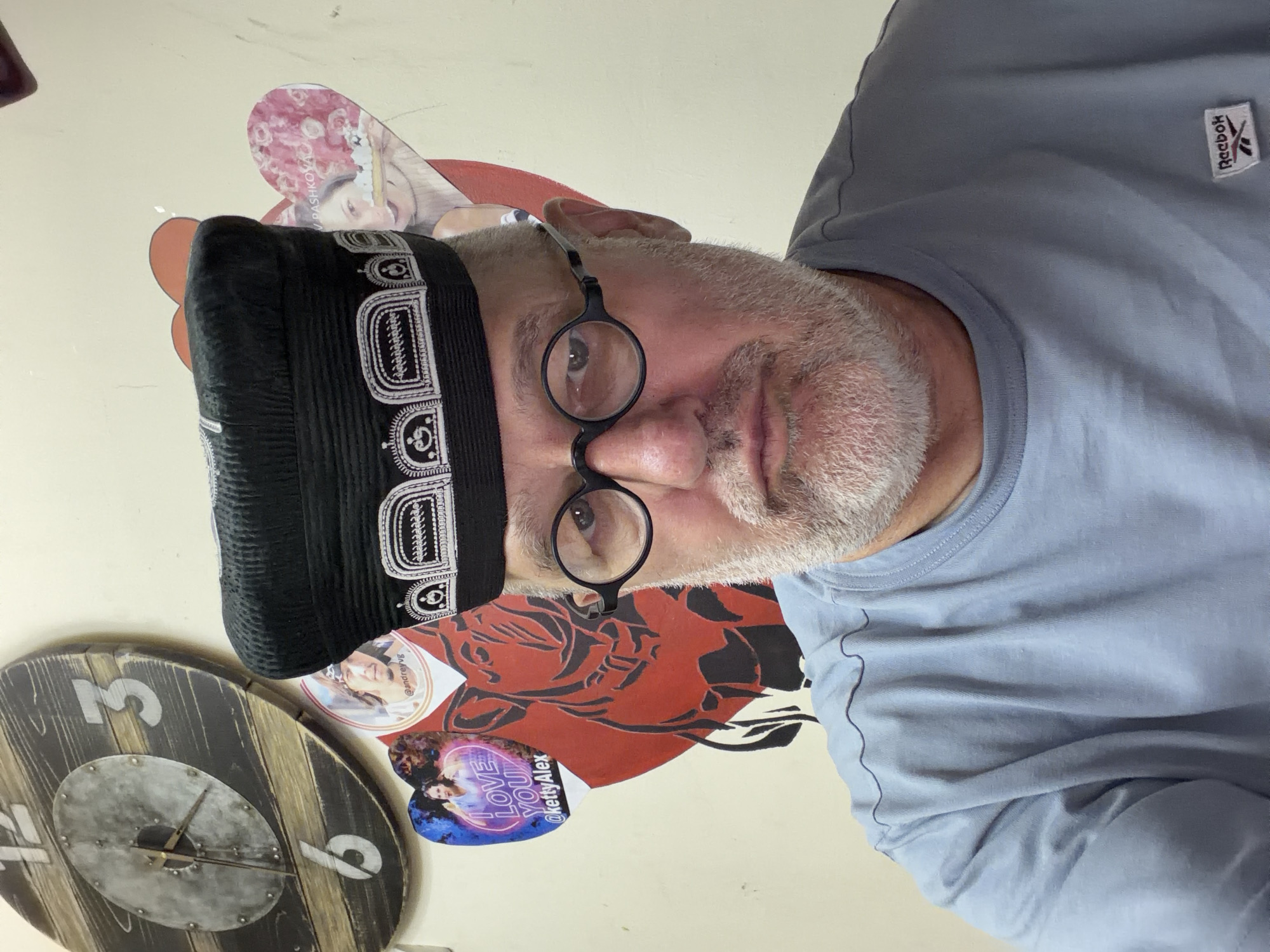 A man with a grey beard and round glasses is looking at the camera and wearing a black cap with white embroidery and a blue t-shirt. Behind him, a wooden clock is hanging on a cream-colored wall. There are also several heart-shaped pictures with images of people and text. One of the pictures has text that says "I LOVE YOU @kettyalex". Another picture has the name "V. PASHKOVA" on it. The man is positioned in the center of the frame.