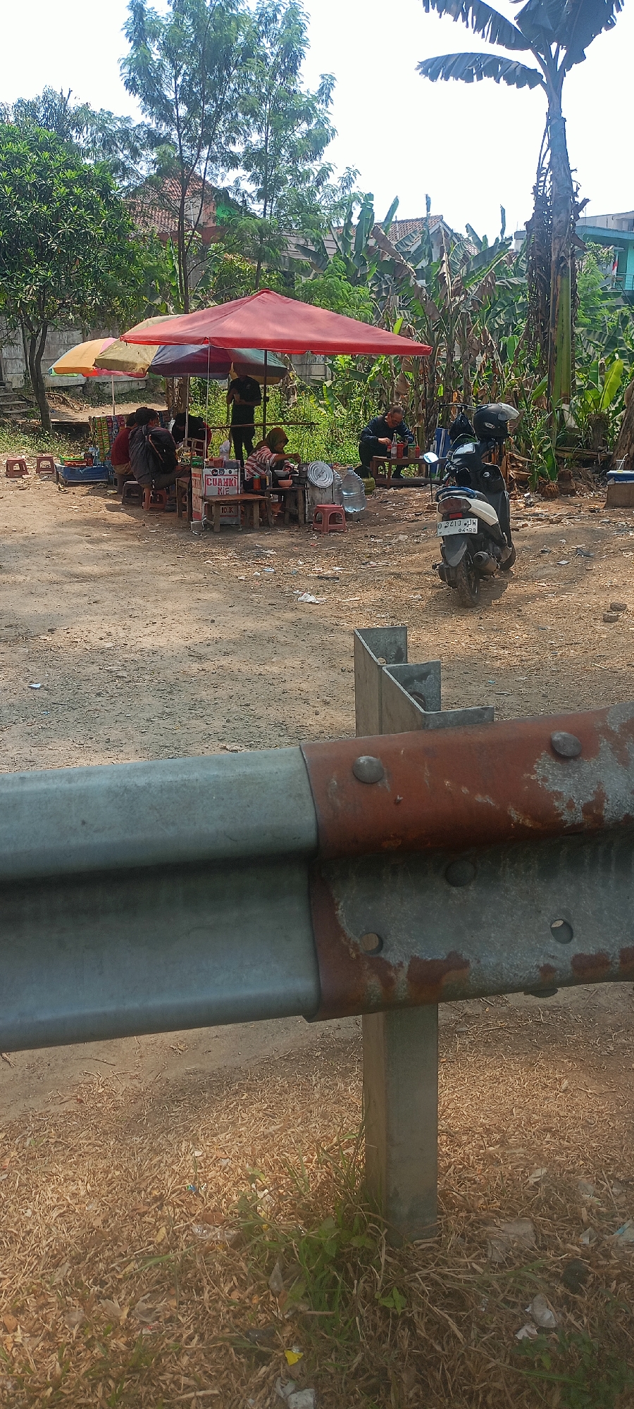 In Kertajaya, Indonesia, this scene captures an outdoor food stall beneath red and yellow umbrellas. Several individuals are seated at small tables, some eating, while others stand. A motorcycle is parked nearby. The ground is dirt, with scattered debris and dry grass. A metal guardrail with signs of rust is visible in the foreground. Lush vegetation, including banana trees, forms the backdrop.
