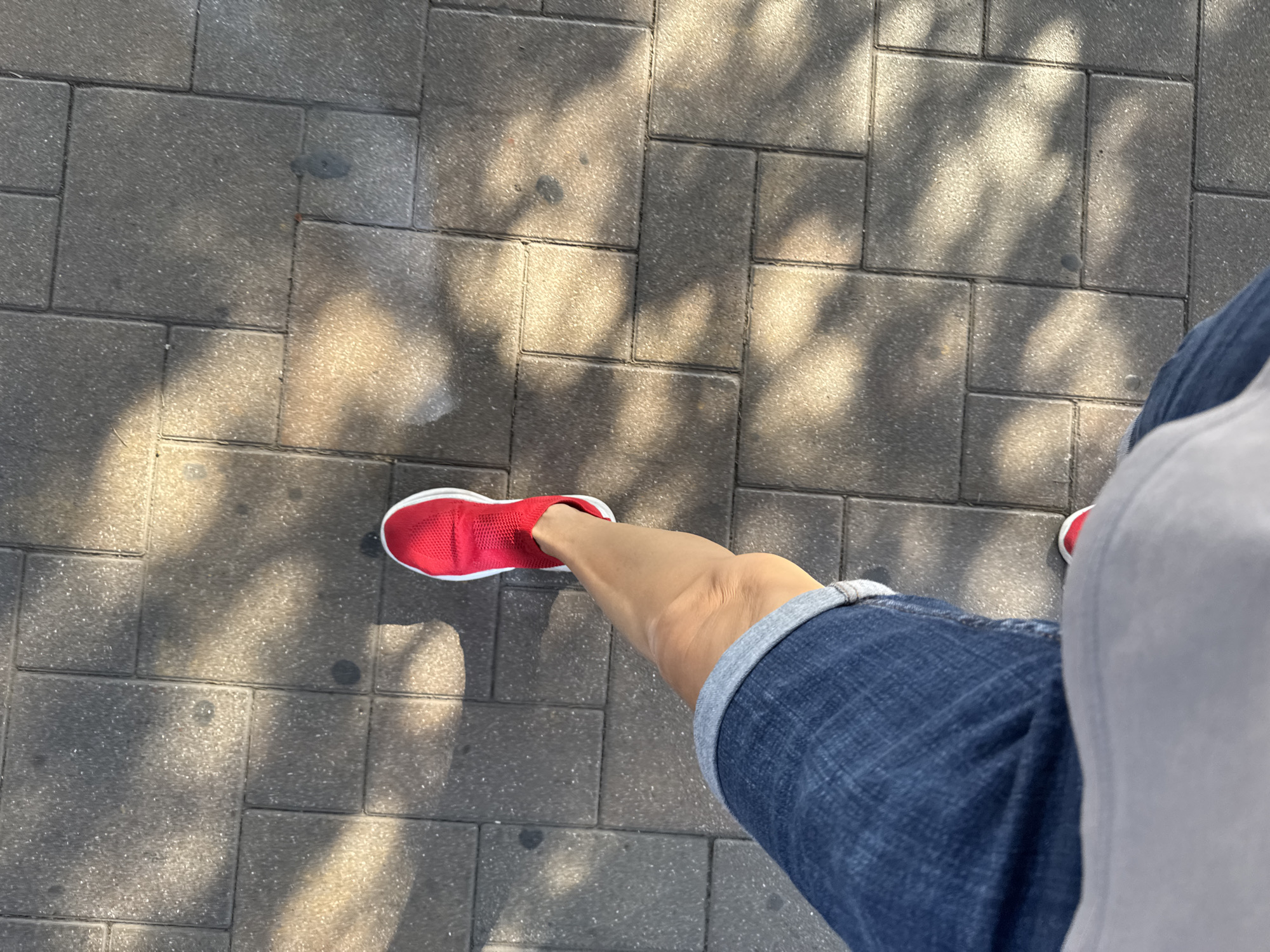 An over-the-shoulder view shows a person walking on a cobblestone sidewalk bathed in dappled sunlight, casting shadows from unseen foliage. The person is wearing blue denim shorts with cuffed hems and a light gray t-shirt. A red athletic shoe is visible on their left foot. The scene is a street in Baku, Azerbaijan.