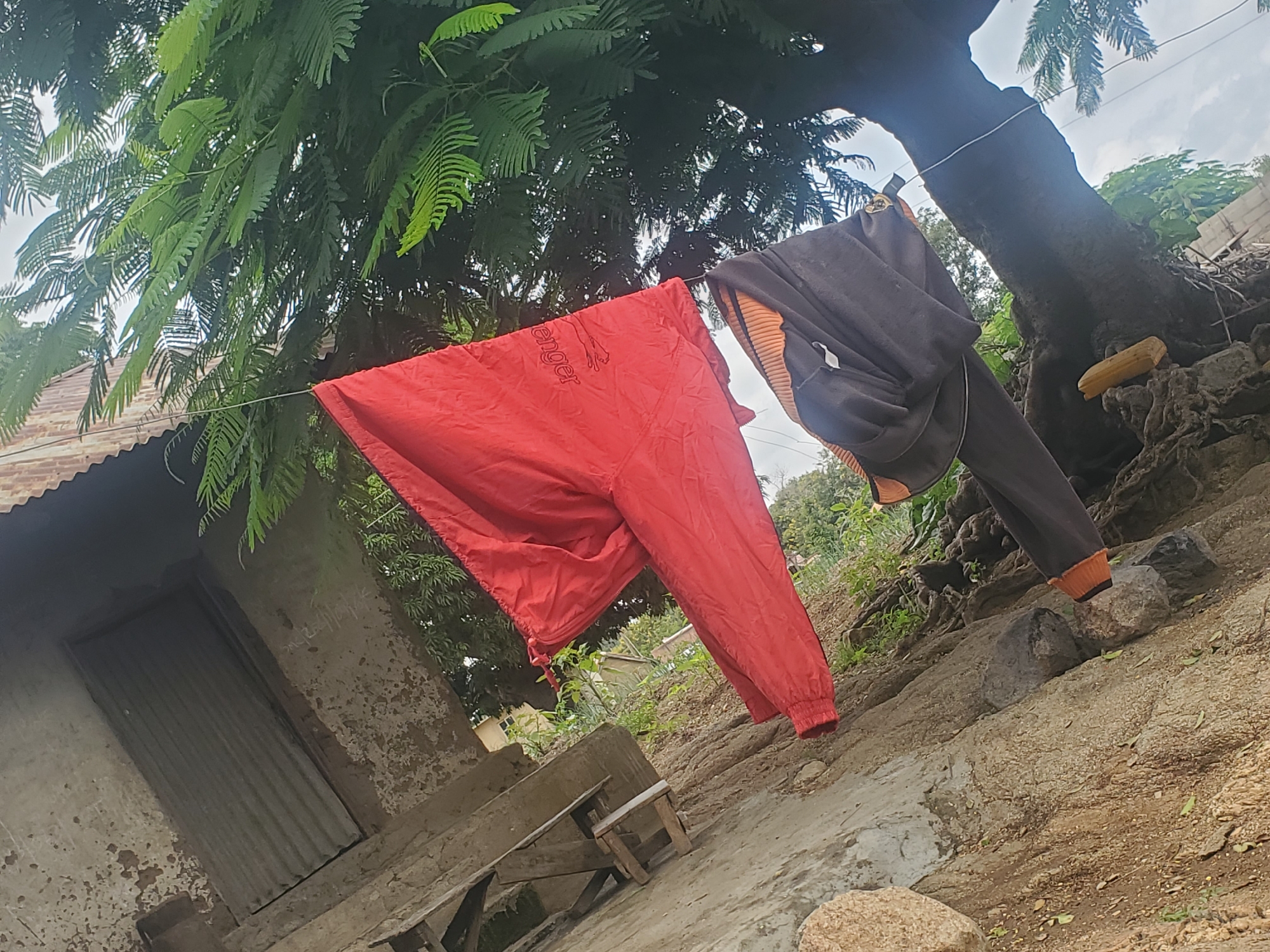 A partially visible red garment and a dark garment with an orange collar are hanging on a clothesline outdoors. The red garment is a jacket with a zipper, and the dark garment appears to be pants. They are hanging from a wire strung between a tree and a structure with a corrugated metal roof. The ground is rocky and uneven, with some vegetation growing around it. The scene is bathed in natural light, suggesting it is daytime.