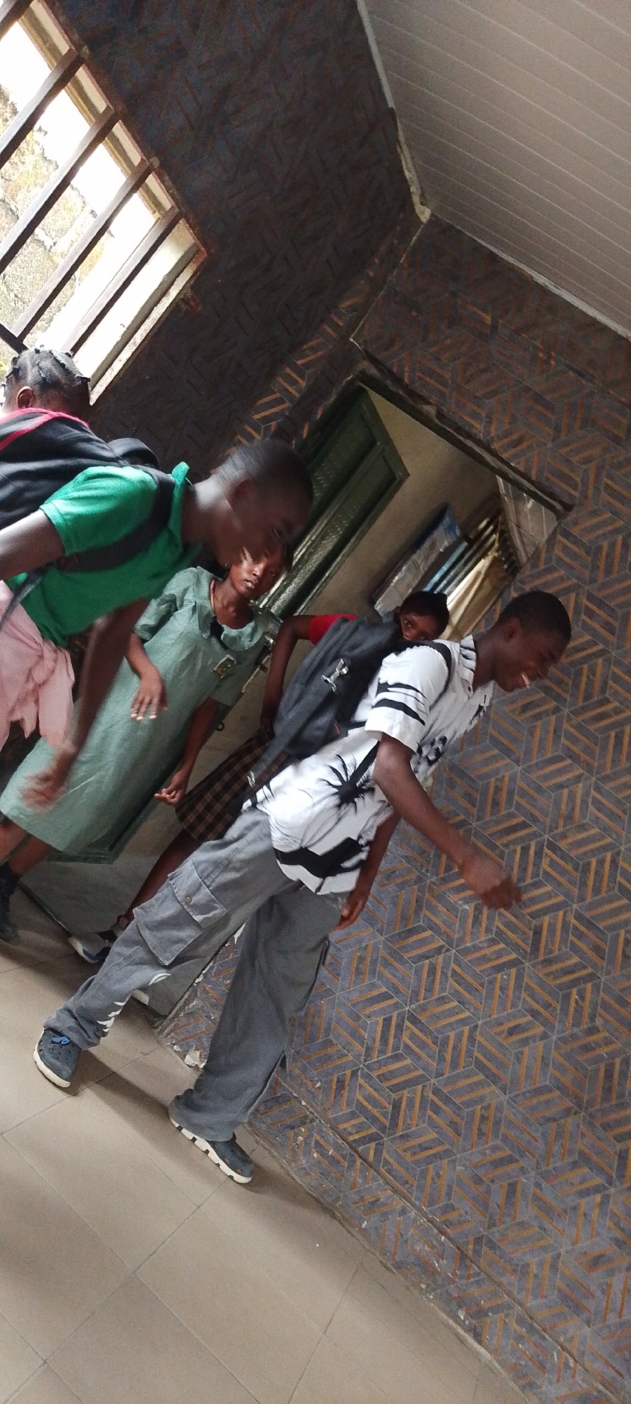 The image depicts a group of children in a corridor within a building, likely a school, in Lokoja, Nigeria. The building features a distinctive tiled floor and a textured wall finish, with natural light entering through a window near the ceiling. The children are wearing varying attire, including school uniforms and casual clothing, and some carry backpacks. Their actions and interactions are not clearly defined, but they appear to be in transit, possibly moving between classes or leaving the building.