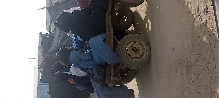 A group of approximately six to seven individuals, predominantly women wearing hijabs and long-sleeved garments, are seated closely together on an improvised, multi-wheeled cart on a dirt road. The cart features a dark, possibly rusted, metal frame and a wooden platform, equipped with at least four small, worn tires with metal rims. The individuals are positioned facing forward or slightly to the right of the frame. In the background, a dense arrangement of makeshift structures is visible under a clear sky, comprising shelters made from corrugated metal sheets and dark tarpaulins. The ground is a light brown, dusty or sandy surface. The scene takes place outdoors during daylight in Khan Yunis, Palestinian Territory.
