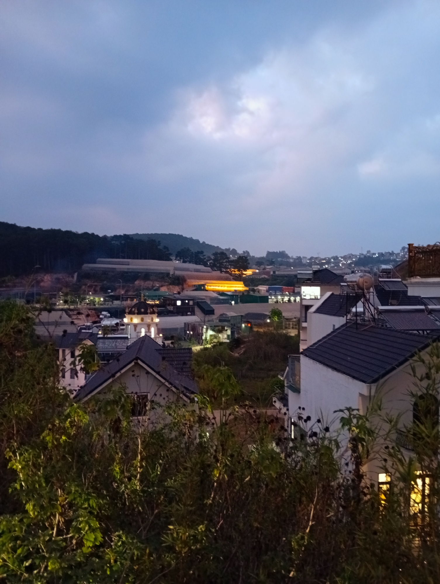 Captured at twilight, the image presents an elevated perspective of a cityscape possibly located in Da Lat, Vietnam, characterized by a mix of residential buildings and agricultural structures. The foreground is dominated by dense shrubbery that obscures a portion of the view. In the midground, several buildings with dark-tiled roofs and white facades are visible, some illuminated by interior lights. Interspersed among the buildings are patches of greenery and small trees. Beyond the buildings, a series of illuminated greenhouses or agricultural structures stretches across the landscape, backed by a dark, tree-covered hill. The sky is overcast with a diffused glow around the horizon, typical of dusk. The light suggests it is evening, with artificial lights becoming more prominent as the natural light fades.
