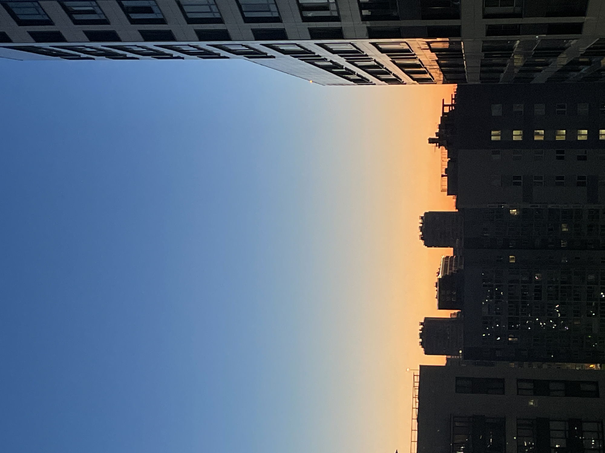 This is a vertical, eye-level photograph taken outdoors in Sinyavino, Russia, at dusk. The image captures an urban landscape with several high-rise buildings silhouetted against a gradient sky that transitions from a deep blue at the top to a bright orange near the horizon. The sky fills the upper two-thirds of the frame, while the buildings occupy the lower third.
On the right side of the image, a tall, modern apartment building dominates the foreground. Its facade is a pale, neutral color, and it features numerous windows arranged in a grid pattern. The building is angled towards the viewer, with its side profile clearly visible. Sunlight reflects off its surface, highlighting the details of its construction.
To the left of this building, and further into the background, a cluster of other apartment buildings is visible. These structures are darker and appear older, with their windows illuminated, suggesting occupancy. They are varied in height and design, creating a dense urban skyline. Some of these buildings have visible balconies and rooftop structures.
The horizon line is low in the frame, marked by the tops of the buildings and the transition between the sky and the cityscape. The overall atmosphere is serene, with the warm colors of the sunset contrasting with the cool tones of the sky and the dark buildings. No people or specific activities are discernible. The photograph appears to be taken from a window or balcony, looking out over the city.