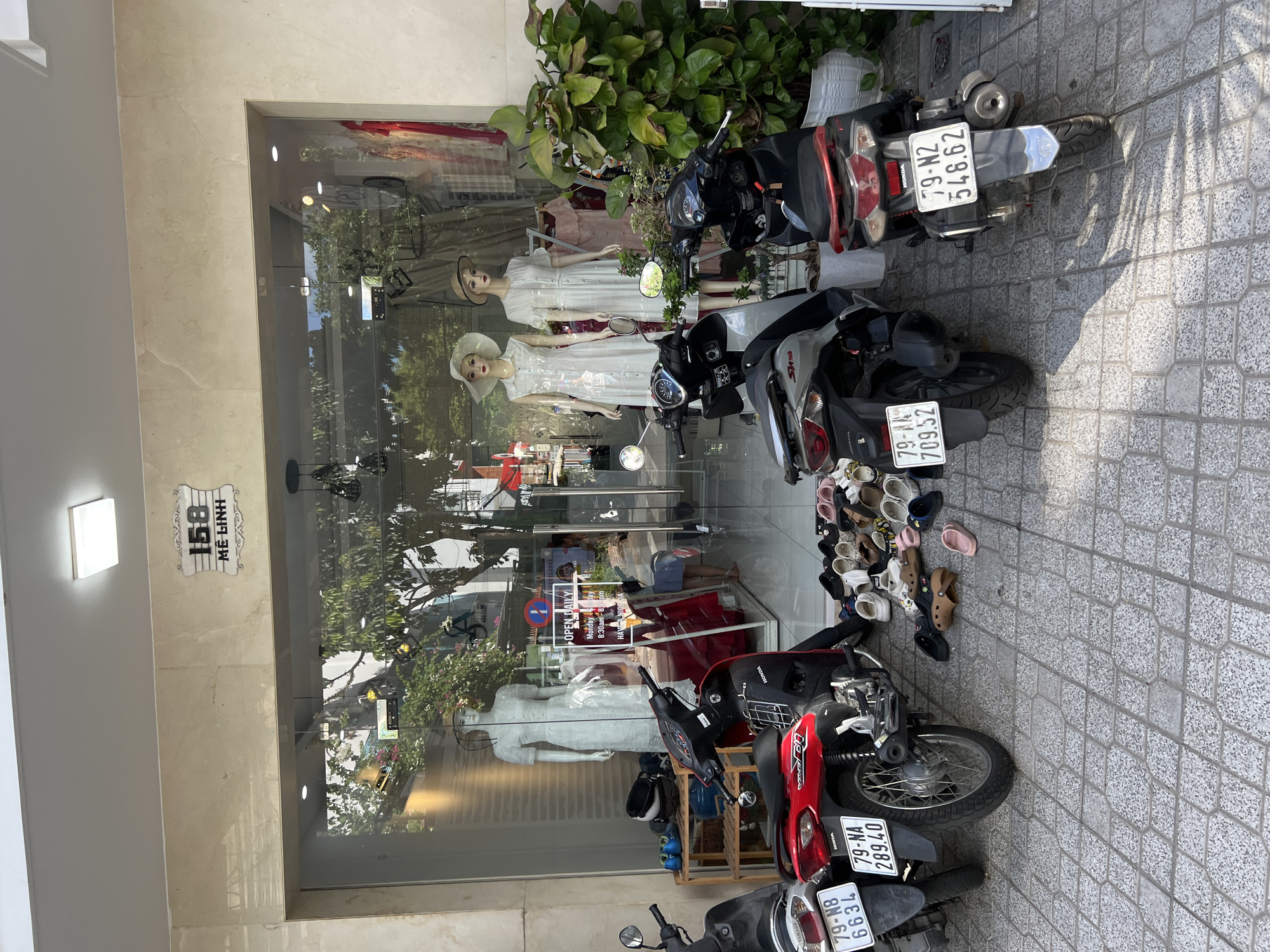The image shows the storefront of "ME LINH 158" in Nha Trang, Vietnam. The store has a large glass facade displaying mannequins in dresses and hats. Several motorcycles are parked in front of the store on a tiled sidewalk, along with a large pile of assorted footwear. A sign on the glass indicates "OPEN DAILY Monday-Sunday 8:30am - 8 pm HAVE A NICE DAY". A person is visible inside the store. Lush green plants are situated to the right of the storefront. The environment appears to be a commercial street on a sunny day.