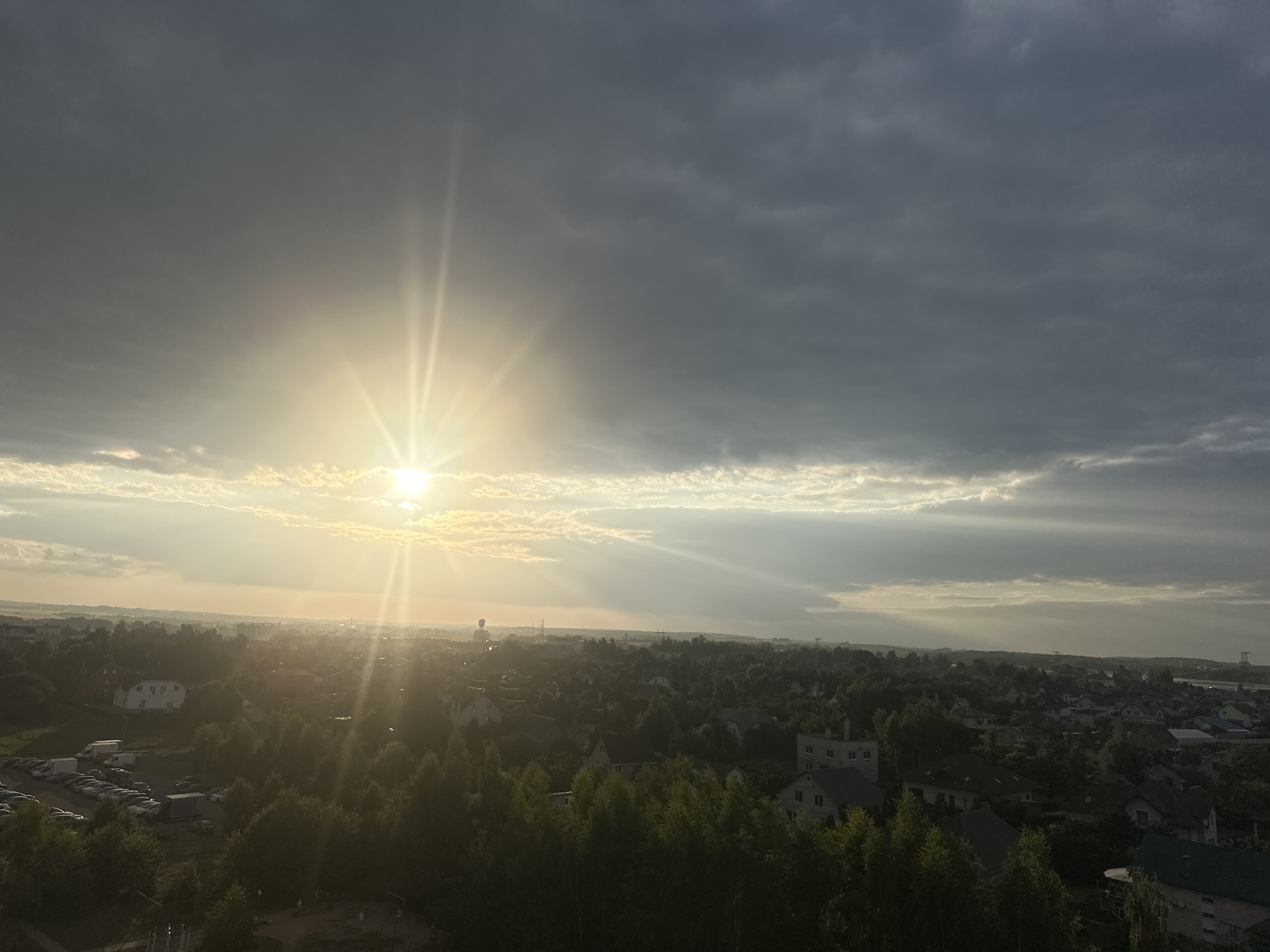 The image captures a wide aerial view of a semi-rural area in the vicinity of the Shchomyslitski Village Council, Belarus, during what appears to be late afternoon or early evening. The sun is setting or has recently set, casting bright rays through a heavily clouded sky. The sun's light illuminates a portion of the clouds from behind, creating a dramatic contrast with the darker, more ominous clouds that dominate the upper half of the frame.
In the foreground and midground, a dense collection of trees forms a dark, textured canopy over a settlement. Visible within this leafy expanse are numerous residential buildings, characterized by varying roof styles and colors, suggesting a mix of older and newer construction. A parking lot filled with numerous vehicles, including cars and trucks, is situated on the left side of the image, adjacent to some of the residences. The terrain appears to be gently rolling, with houses nestled amongst the trees and on slight inclines.
The background shows a more distant urban or industrial landscape, indicated by taller structures and a clearer horizon. The overall atmosphere is one of diffused light and encroaching dusk, with the sun's rays piercing through gaps in the clouds, casting beams that extend across the landscape and create a somewhat hazy effect over the distant scenery. No people or specific actions are discernible in the image. The scene depicts a quiet residential area on the outskirts of a populated region, under a dramatic sky.
