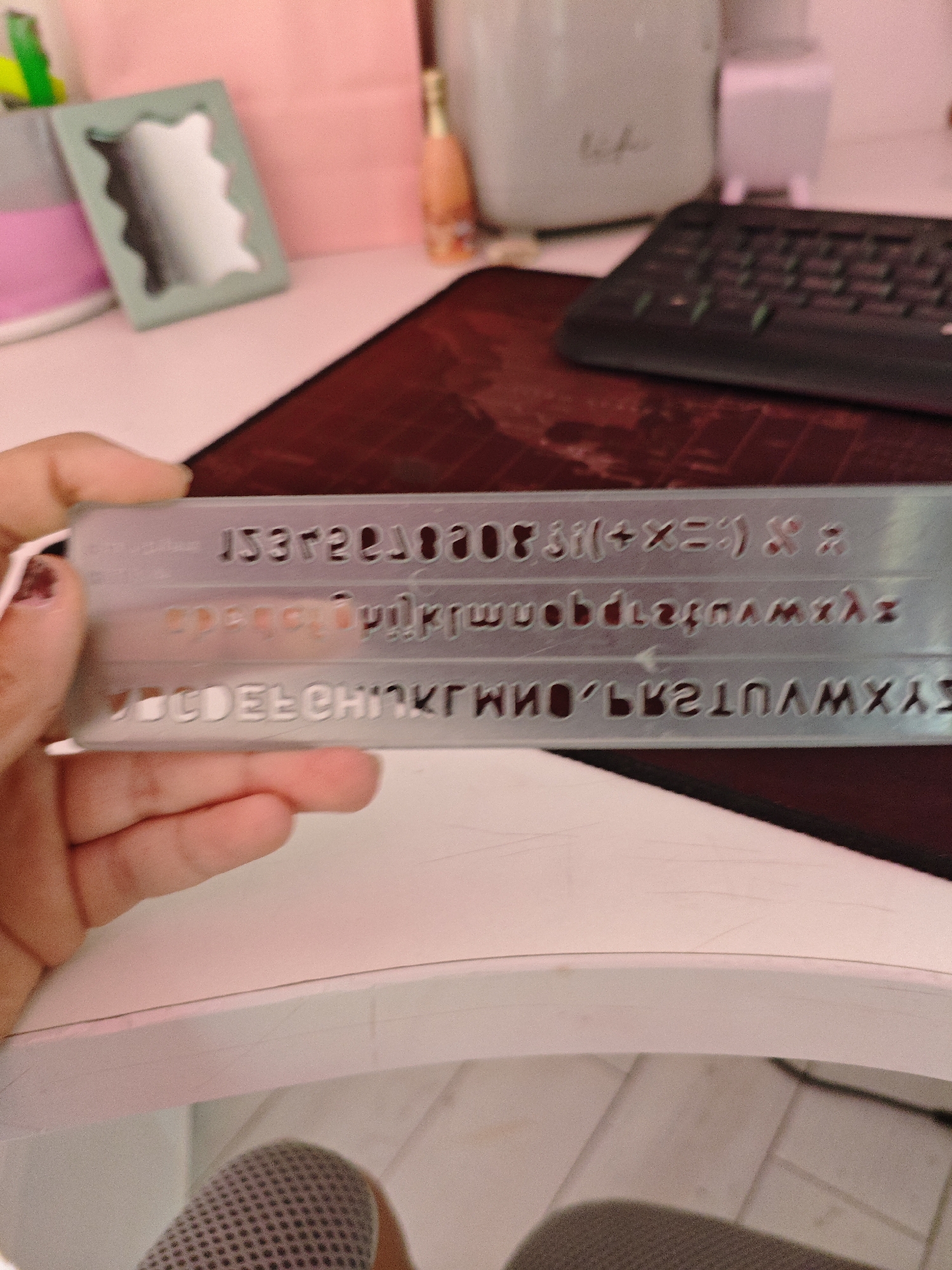 A person's hand is holding a stencil with rows of cut-out letters and symbols, including numbers, punctuation, and the Greek letter lambda. The stencil is silver and translucent. In the background, a keyboard is visible on a dark red mousepad. Other blurred items include what appears to be a small decorative mirror, a pink folder, a bottle, and a white container. The scene is lit with a pinkish light, suggesting an indoor environment. The exact location in Трудовское сельское поселение, Ukraine cannot be determined from the visual information provided.