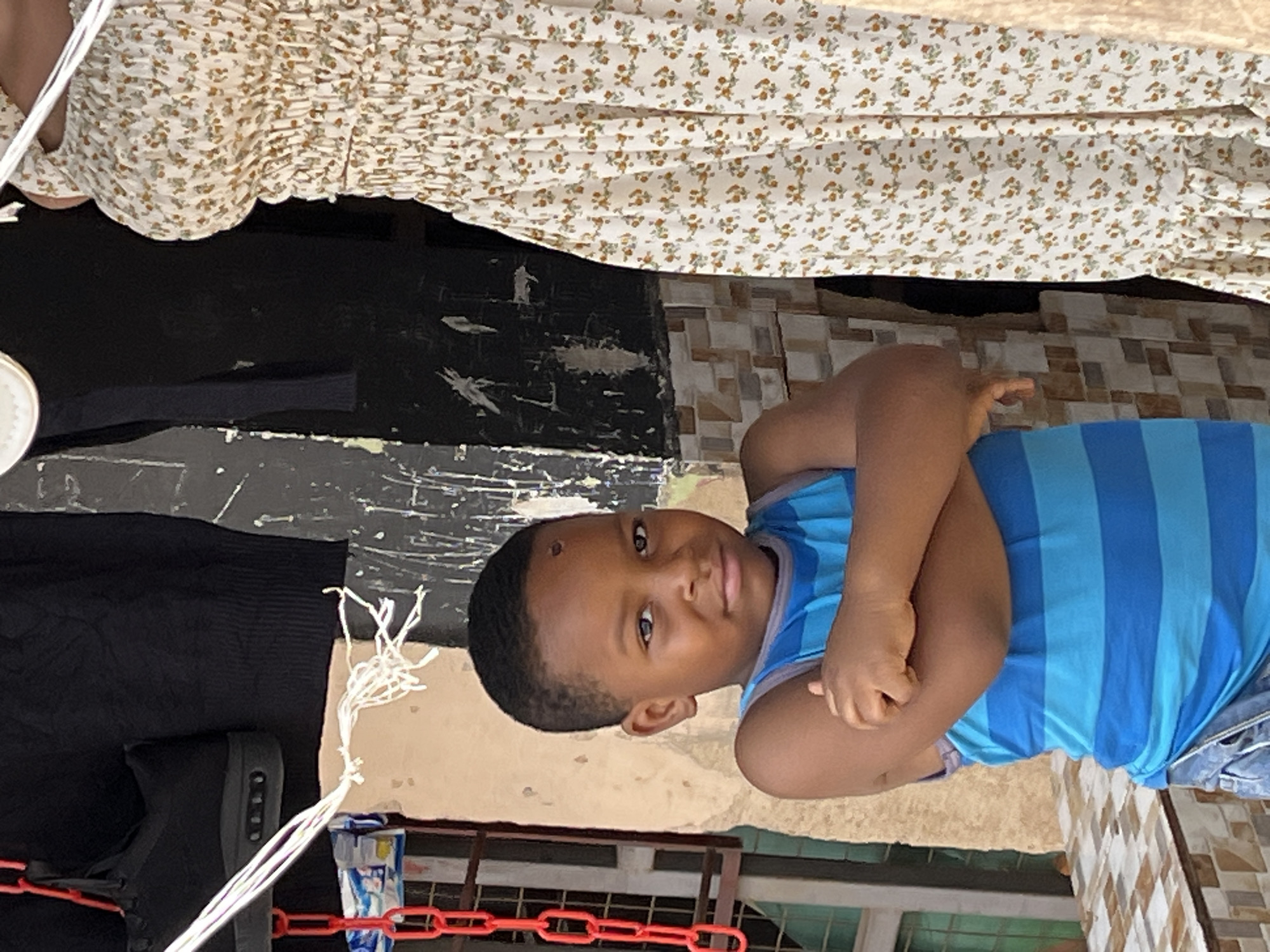 A young boy in a blue striped tank top lies on the ground, arms crossed. He has dark hair and skin. He is on what appears to be a tiled outdoor surface. Above him is a person in a light-colored floral print dress. To the boy's left is a black object and a red plastic chain. A city location cannot be determined.