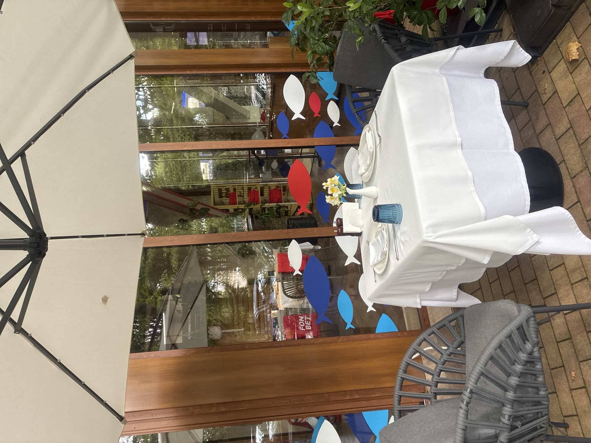 This image depicts an outdoor seating area at a restaurant, likely in Sochi, Russia, given the context provided. A white umbrella is partially visible overhead, casting shade over a table set for dining. The table is covered with a white tablecloth, adorned with place settings that include plates, silverware, and a blue glass. A small vase with yellow flowers sits on the table, adding a touch of decoration.
The restaurant's interior is visible through large glass windows, reflecting the outdoor environment and revealing glimpses of tables and chairs inside. Decorative elements, such as cutouts of blue, white, and red fish, are affixed to the exterior of the glass, suggesting a nautical or marine theme. One prominent red cutout displays the text "FON BET."
Two chairs are positioned at the table. One is a gray wicker chair with a padded seat, while the other appears to be a dark-colored wicker chair, partially obscured. The seating area is paved with brick tiles, and fallen autumn leaves are scattered on the ground. Lush green plants are visible to the right, adding to the outdoor ambiance. The overall scene suggests a tranquil setting for a meal.