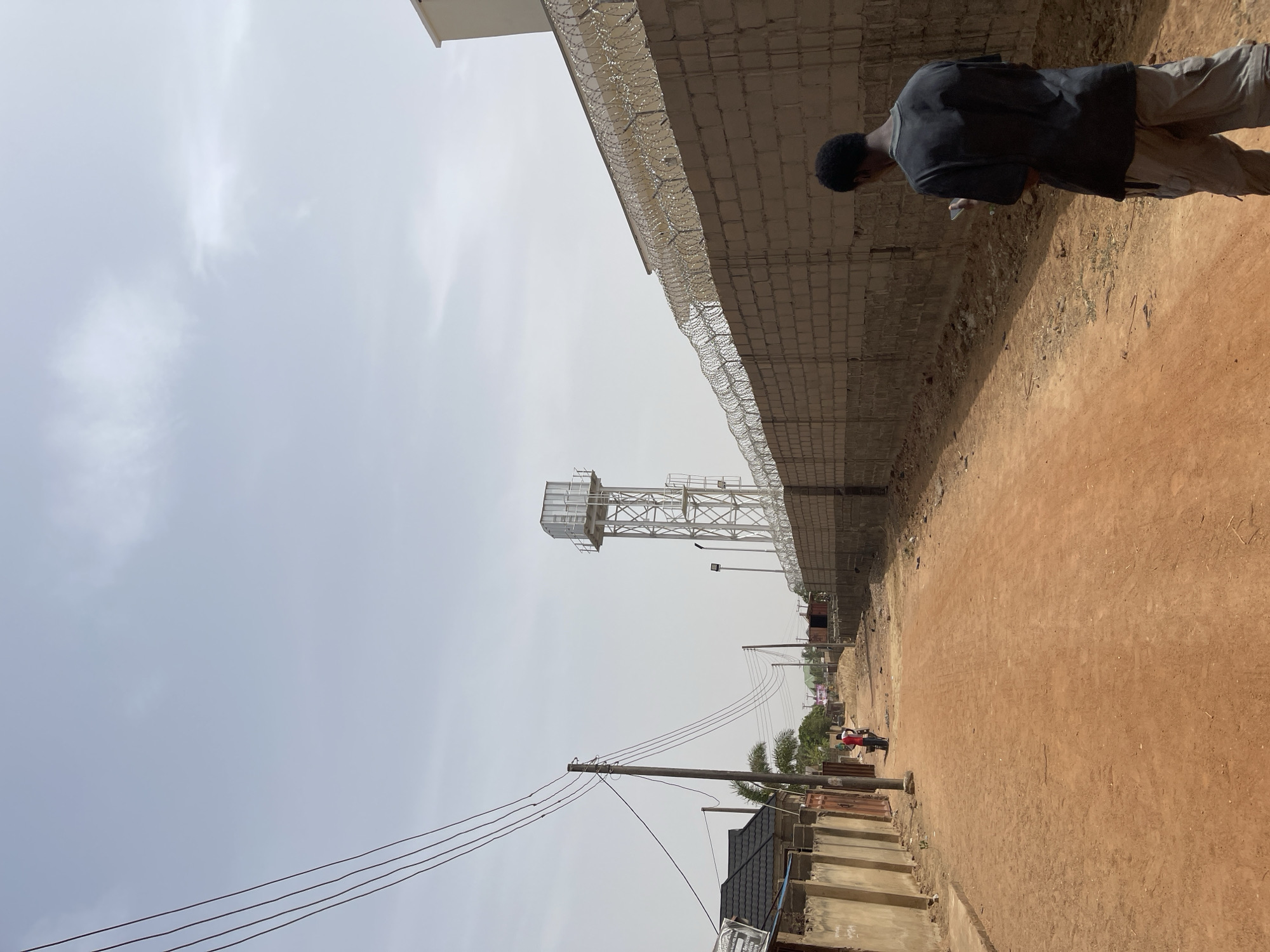 The scene is an unpaved road in Sabon Tasha, Nigeria. A man in a dark shirt and light pants stands near a high, concrete block wall topped with razor wire. The road is dusty and extends into the distance, flanked by the wall on one side and low buildings and fencing on the other. Utility poles and wires run along the left side of the road, leading to a water tower in the distance. Other people are visible further down the road.