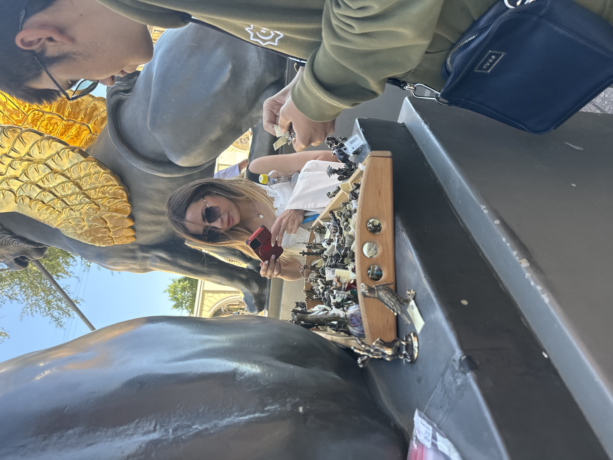 In Saint Petersburg, Russia, near the historic Anichkov Bridge, two individuals are browsing a display of small metal figurines. The woman, wearing sunglasses and a white top, is holding a red smartphone, appearing to take a photo. The man, in a green hoodie, is examining a figurine in his hands. The figurines are arranged in a wooden tray, showcasing various small animals and figures. In the background, a large statue of a horse with golden wings, a prominent feature of the bridge, is visible. The scene is set against a clear blue sky, suggesting a pleasant day.