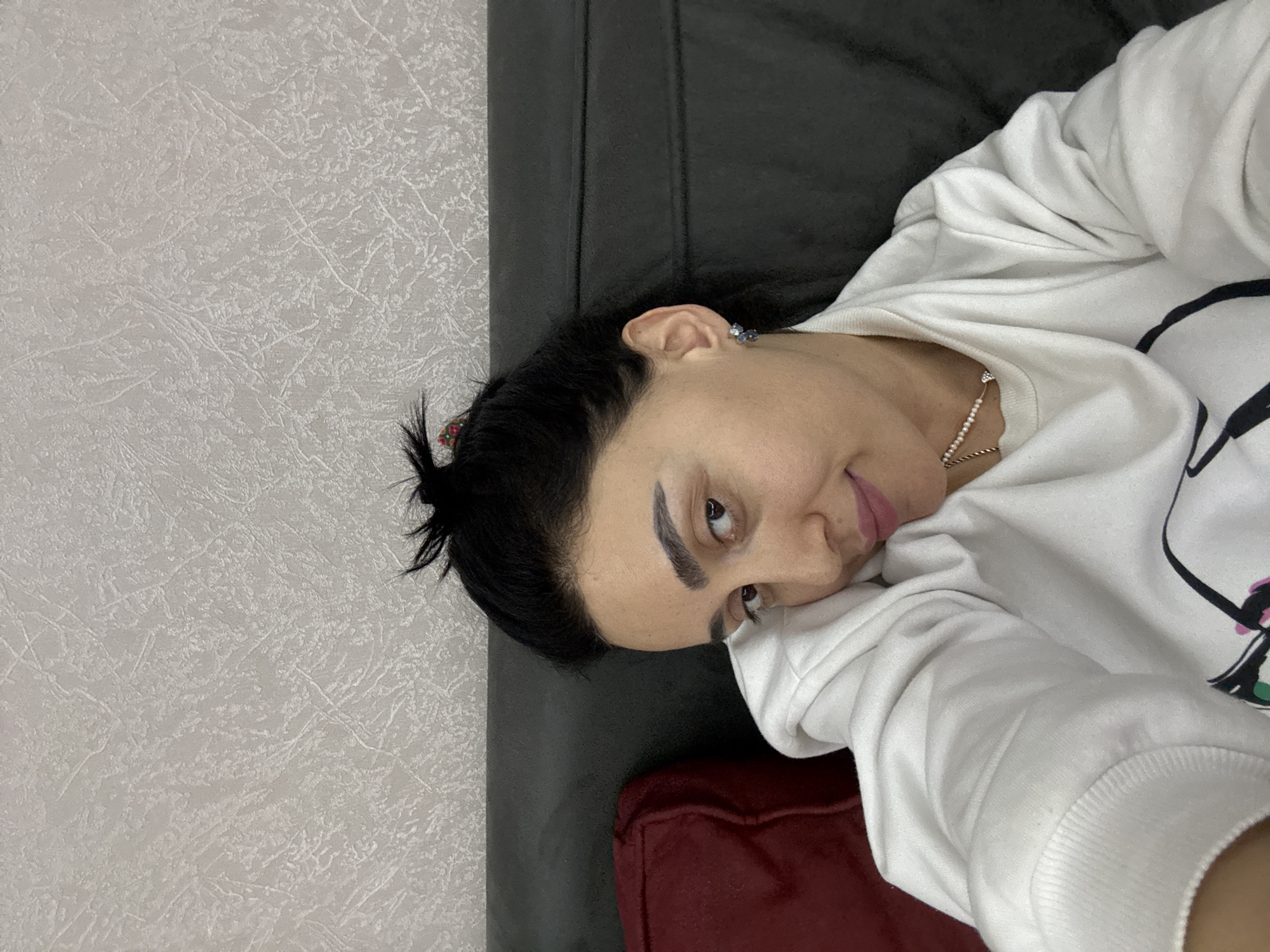 A woman with dark hair and eyebrows is lying on a gray sofa. She is wearing a white sweatshirt and a necklace. She is in a room with a textured white wall. The location is likely Tbilisi, Georgia.