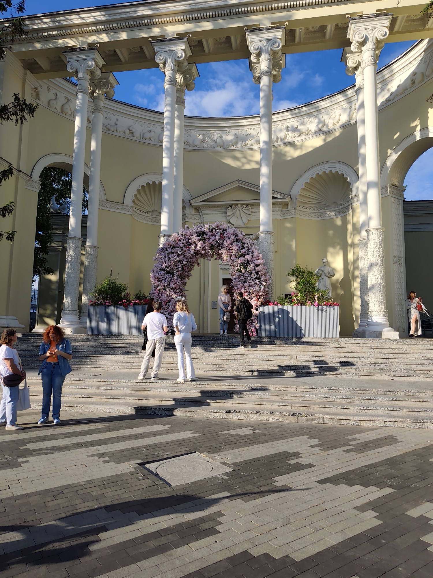 The image captures a scene at VDNKh (Exhibition of Achievements of National Economy) in Moscow, Russia. The primary focus is on a grand, classical building with white columns and arches, typical of Soviet architecture. The building is adorned with animal relief sculpture and has a floral archway installed at its entrance.
Several people are present. A group of three women are standing in the lower left, while a few people are interacting near the floral arch. These individuals are engaged in conversation or taking photographs.
The environment includes a paved square in the foreground, made of patterned bricks. Stairs lead up to the building. Shadows cast by the sun suggest it's daytime, with clear skies visible through the arches of the building. The overall atmosphere is that of a public space, likely a popular spot for visitors and photography.