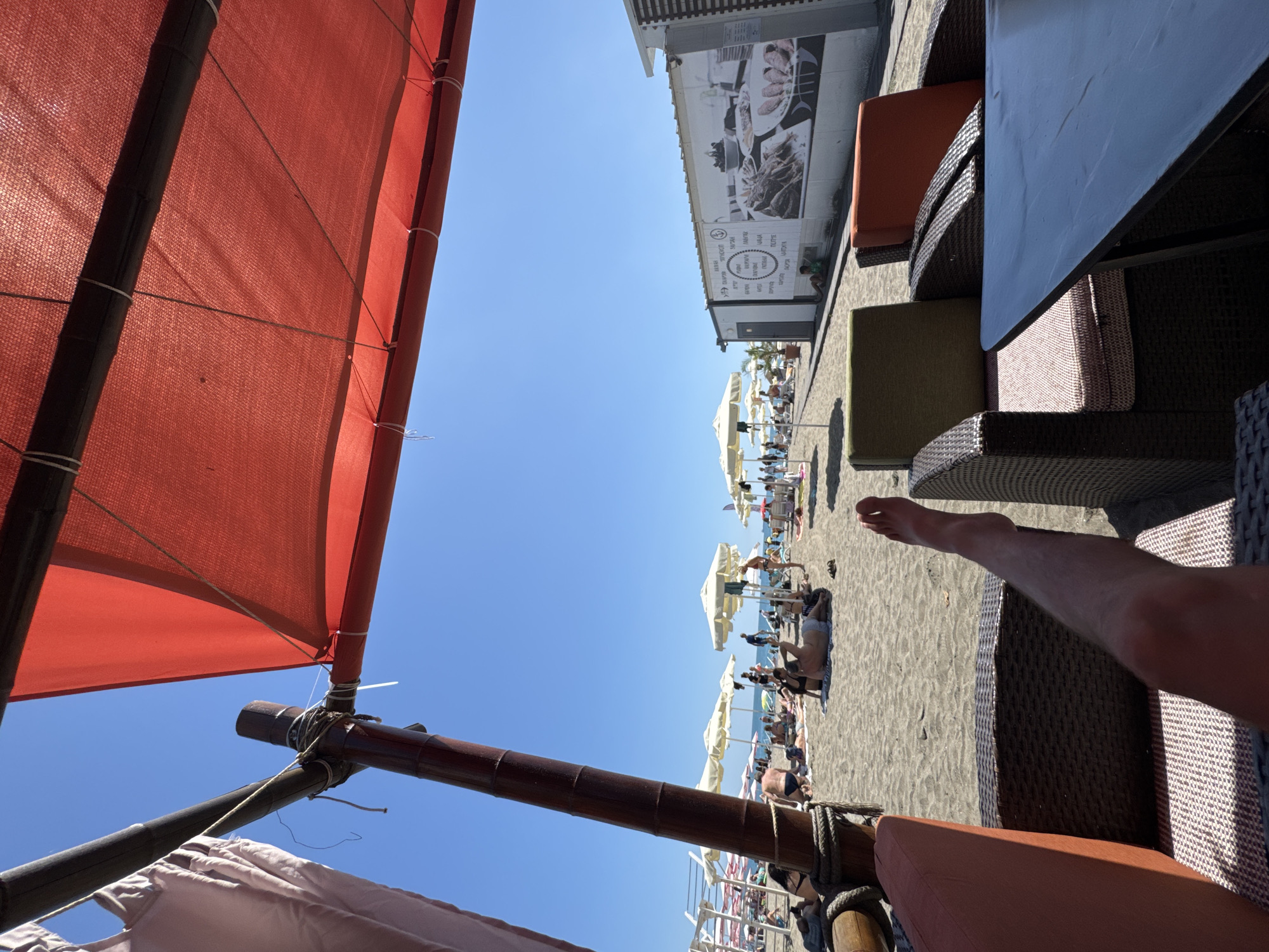 A first-person perspective shot on a sandy beach in Burgas, Bulgaria, shows a person's leg extended towards a table. The leg is bare from mid-thigh down, indicating a relaxed posture. In the foreground, wicker chairs with colorful cushions flank a dark table, partially obscuring the view of the beach. The beach itself is populated with people sunbathing and sitting under umbrellas. The sea and a clear blue sky are visible in the background, along with a white building featuring a large mural of food. The scene suggests a leisurely day at a beachside establishment.
