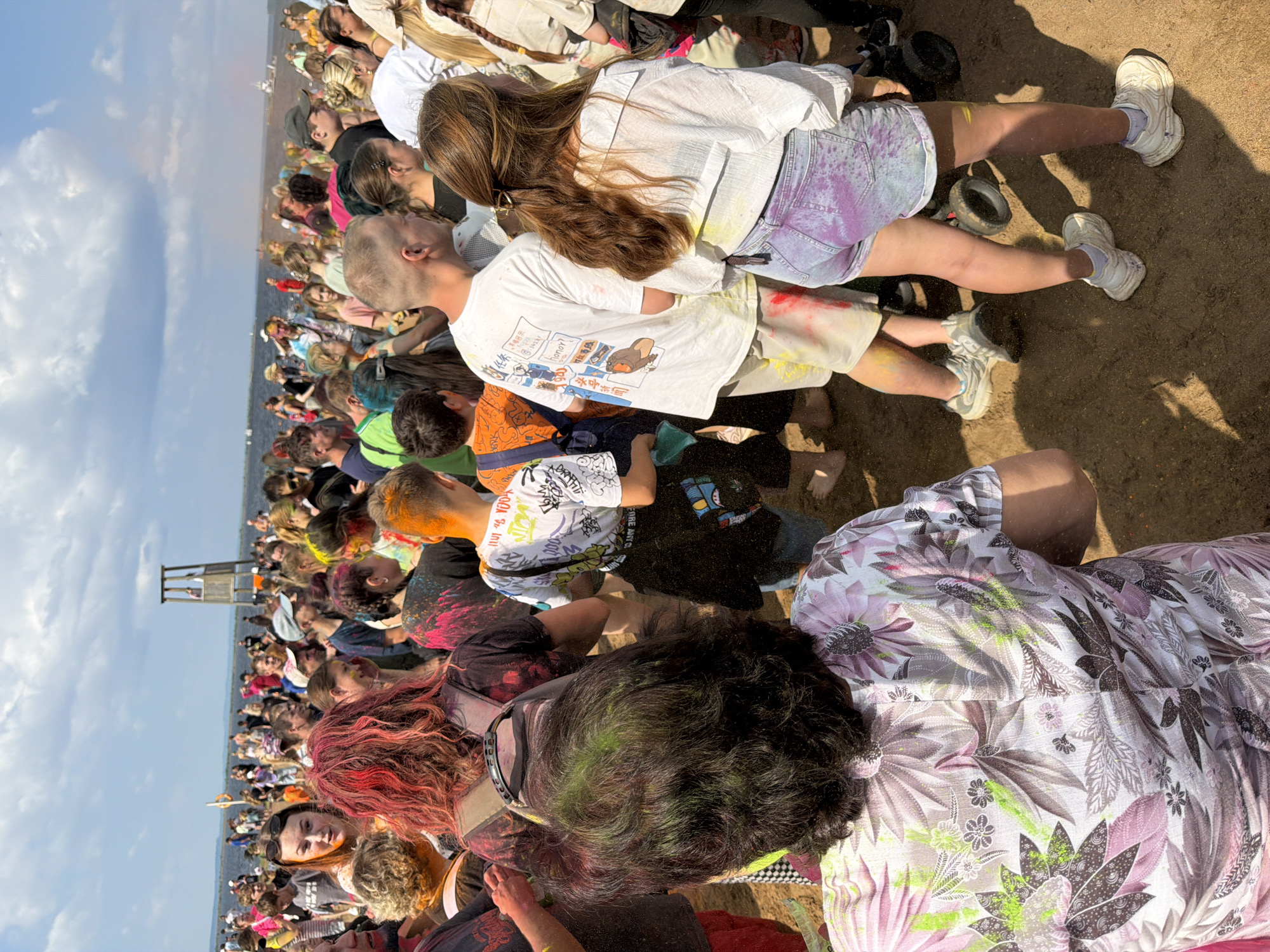 The image captures a crowd of people participating in a color festival, likely near the Kama River in Chaikovsky, Russia. The participants are covered in vibrant powder paint, indicative of the Holi festival or a similar event. In the background, a wooden tower stands by the water, and a hazy rainbow effect is visible in the sky, possibly due to airborne colored powder. The ground is sandy and littered with fallen powder, further suggesting the festive nature of the gathering. The people, mostly young adults, are dressed casually in t-shirts, shorts, and some are wearing backpacks. The atmosphere appears lively and celebratory, with a sense of communal engagement in the colorful activities.