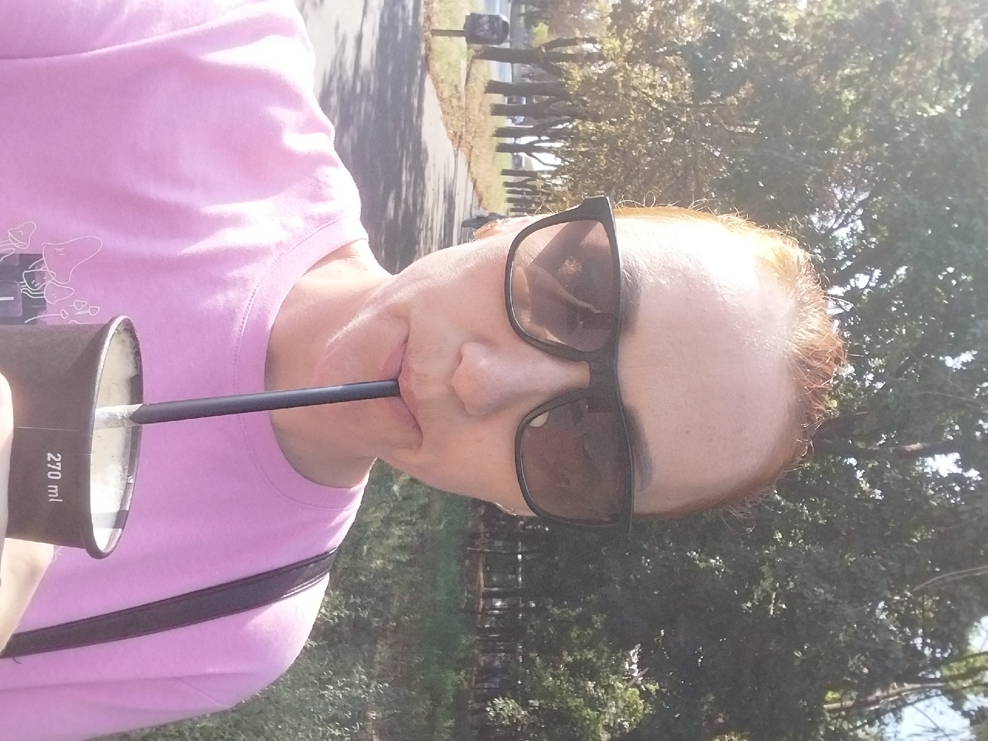 The image depicts a woman with reddish-blonde hair, wearing large black sunglasses and a pink t-shirt, drinking from a paper cup with a straw. She is outdoors, likely in a park or along a paved path lined with trees. The background suggests a sunny day with dappled sunlight filtering through the leaves. A mailbox is visible on the left side of the path, and the overall environment appears to be urban or suburban. The cup has "270 ml" printed on it. There are no visible indications of the specific location within the city of Dnipro, Ukraine.