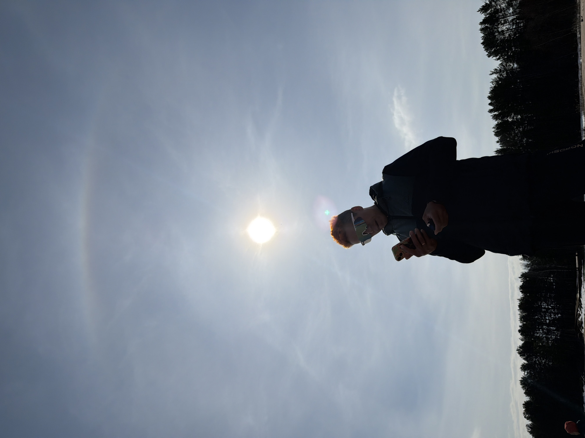 The image depicts a person standing outdoors against a bright sky with the sun visible. The person is holding a mobile phone and appears to be looking at it. The person has orange-red hair and is wearing sunglasses. Dark colored trees line the horizon. A circular halo around the sun is visible in the sky. The location might be the city of Chaikovsky, Russia.