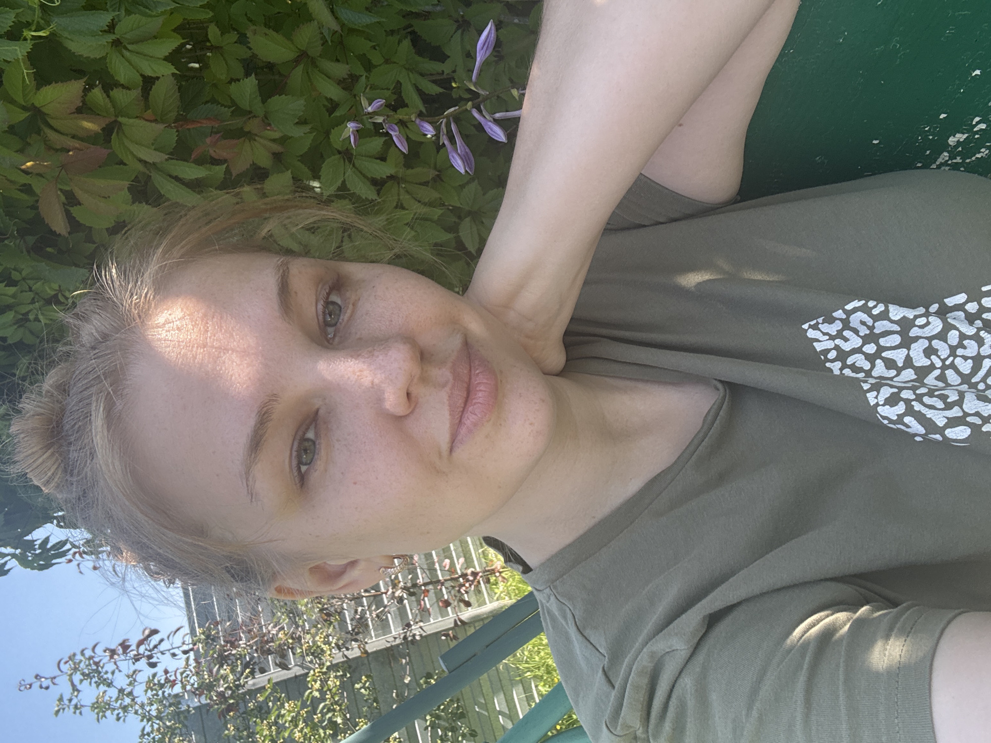A selfie is taken from a low angle of a young woman with blonde hair pulled up in a bun, looking at the camera. She has fair skin with freckles and is wearing a green t-shirt with a white leopard print design on it. Her right arm is resting on her neck. She is outdoors, with lush green foliage and a clear blue sky in the background. The woman appears to be sitting on a green bench or swing. In the background, to the left, there is a light-colored fence and some trees. To the right, there is a plant with purple flowers. The scene suggests a warm, sunny day.