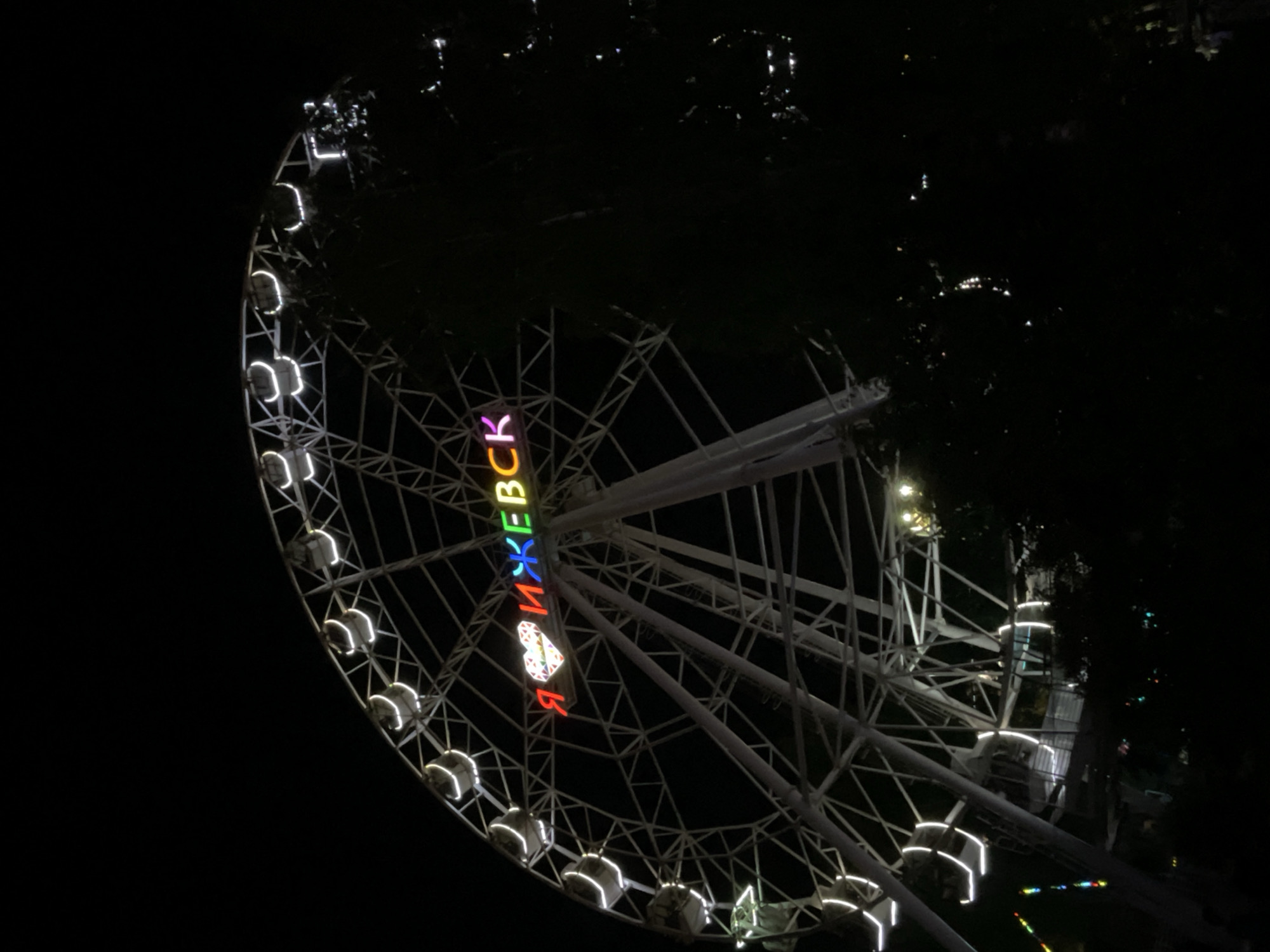 The image shows a Ferris wheel at night in Izhevsk, Russia. The Ferris wheel is white and illuminated with lights. Some of the lights are white, while others are colored and spell out text. The text visible on the Ferris wheel appears to be "Я ИЖЕВСК," which translates to "I Izhevsk." The surrounding area is dark, suggesting it is nighttime. There are trees visible in the background, and some distant lights can be seen, indicating a city or town setting. The Ferris wheel is the primary subject of the image. The scene appears to be at an amusement park or fairground. The lighting creates a festive and somewhat magical atmosphere.