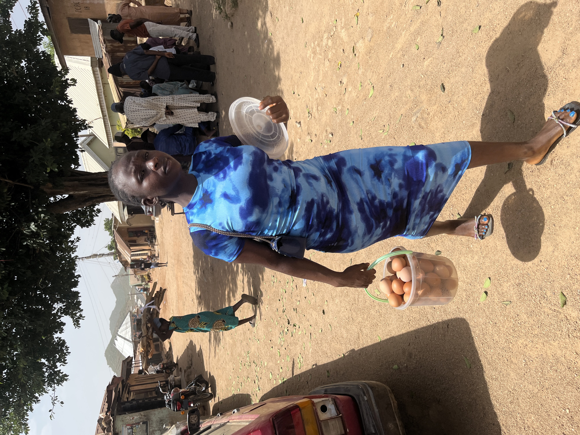 A woman stands on a sun-drenched, dusty ground surface, holding a plastic container filled with eggs and a lid in each hand. She is wearing a blue dress and sandals with bedazzled straps. The environment includes a partial view of a car in the lower left corner, and a group of people gathered near buildings in the background, along with an additional figure carrying something on their head. The setting suggests a location in Jos, Nigeria due to the style of buildings, clothing, and the arid landscape.