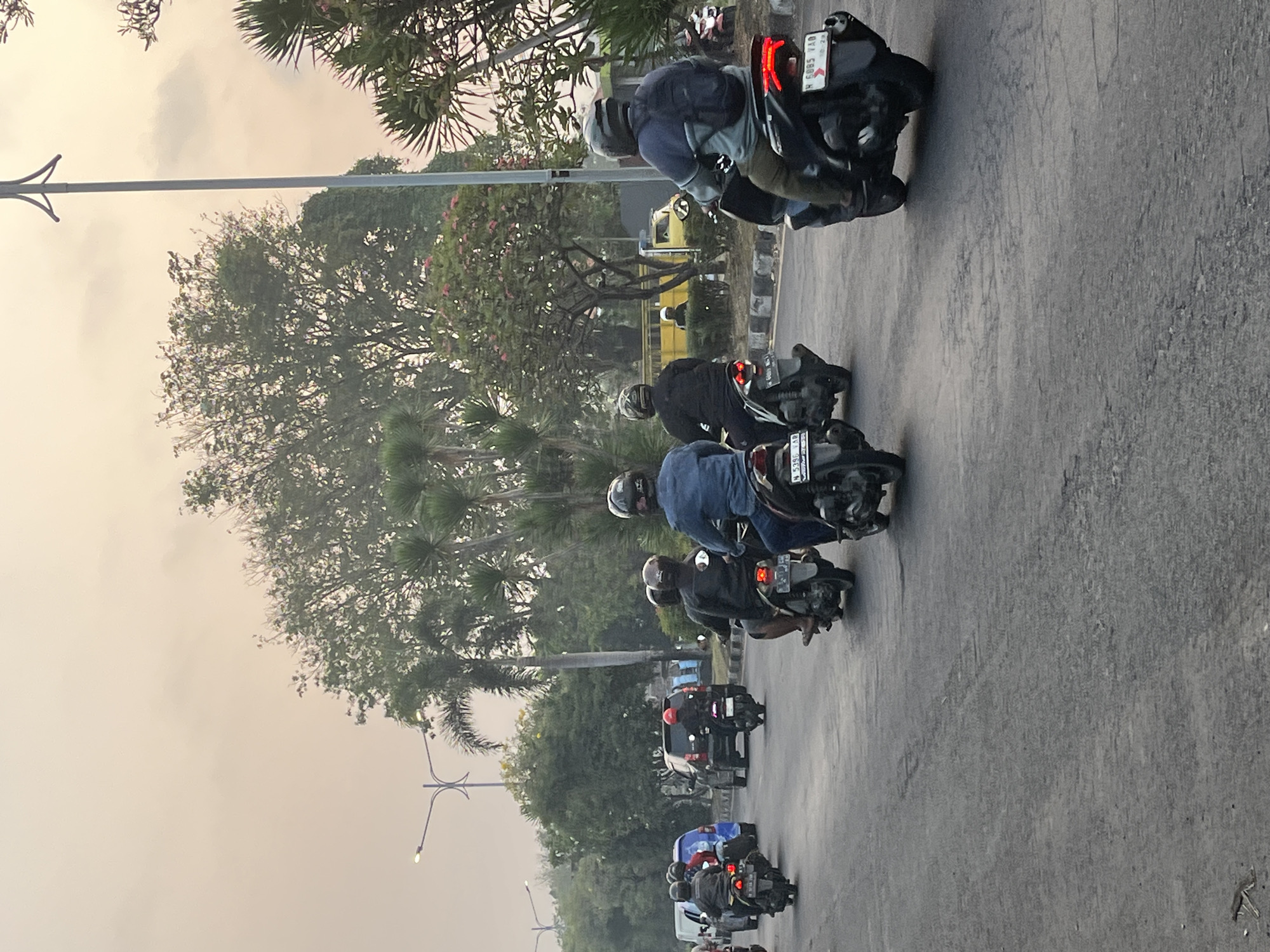 The image depicts a street scene in Kludan, Indonesia, likely during the late afternoon or early evening, indicated by the soft, diffused light and overcast sky. Several motorcycles are traveling on a paved road, with some riders wearing helmets and backpacks. The road appears to be a main thoroughfare, with cars and a yellow truck visible in the background. Trees and lush vegetation line the sides of the road, suggesting a somewhat rural or suburban setting within Kludan. A streetlamp is also visible, further contributing to the urban infrastructure. The overall atmosphere is one of everyday life, with people commuting or traveling on their motorcycles.