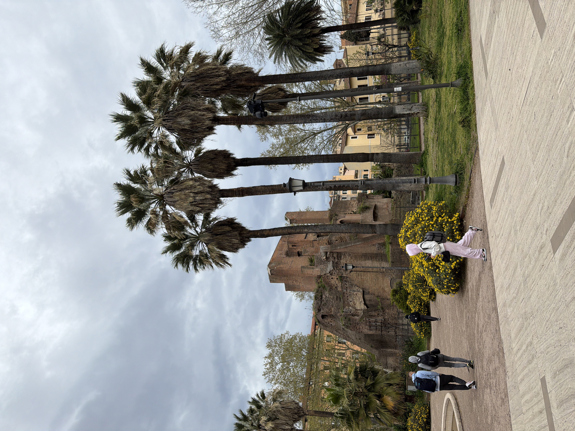 The image shows a street scene in Rome, Italy, lined with tall palm trees. In the background is a brick building or ancient structure, likely part of the ruins in the city center. Several people are walking along the paved walkway, including a person wearing a pink outfit, and others in casual clothing. Street lamps are interspersed among the trees. The sky is overcast. Yellow flowers are present in the landscaping.
