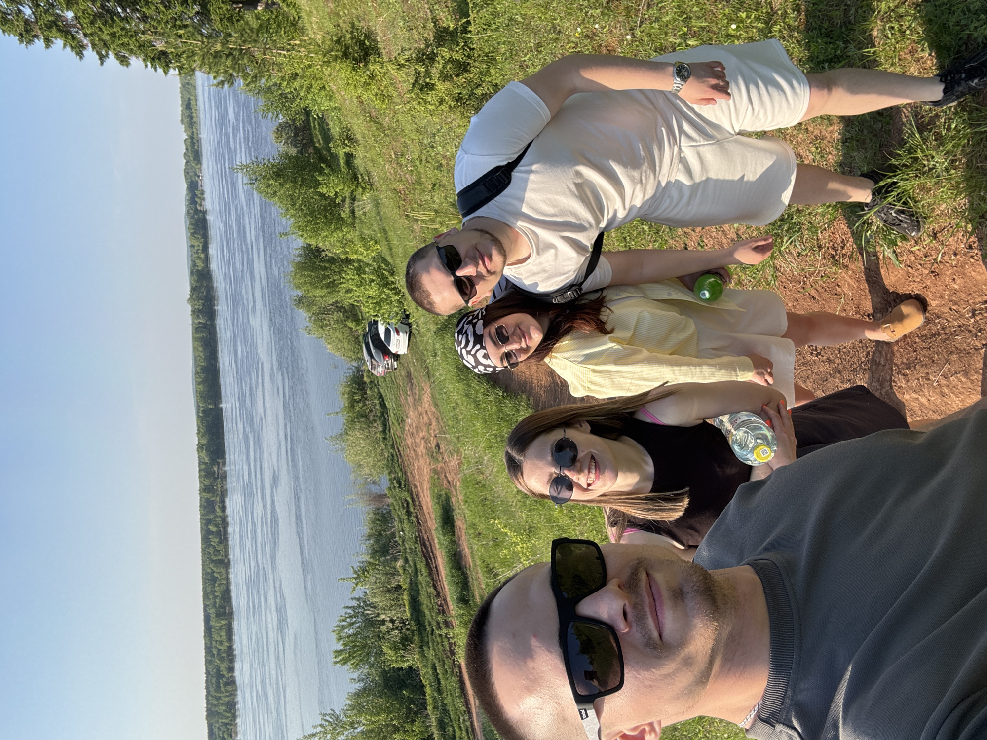 Four people are captured in a selfie taken outdoors. The background features a wide river bordered by green vegetation under a clear sky. The person taking the selfie is at the bottom of the frame, wearing sunglasses and a grey shirt. Above this individual is a woman with long brown hair, wearing sunglasses and a black top, holding a water bottle. Above her is another woman wearing a yellow shirt, sunglasses, and a patterned head covering. At the top of the frame is a man wearing sunglasses, a white t-shirt, and white shorts. The location is in Ольховка, Russia, based on visual cues.
