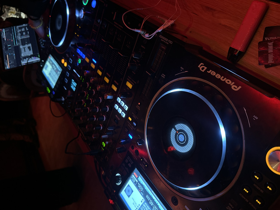 The image displays a close-up, high-angle shot of a DJ setup, likely in a club or bar environment, characterized by dim lighting and vibrant glowing controls. The main focus is on a professional DJ mixer and two Pioneer DJ CDJ-2000NXS2 media players, arranged on a dark wooden surface.
On the left side of the frame, a hand is visible near a tablet that is displaying a complex interface with waveforms and other music production or performance data, suggesting active engagement with the DJ equipment. Below the tablet, one of the CDJ players features a bright screen showing track information, including a waveform, tempo (130.0 BPM), elapsed time ("0:52:46.226"), and a track status ("-0.05 856"). The jog wheel of this player is illuminated with a bright ring.
In the center, the DJ mixer has numerous illuminated buttons and knobs, glowing in blue, green, and yellow, indicating various active functions. Cables from headphones are visible connecting to the mixer or one of the players.
To the right, another Pioneer DJ CDJ-2000NXS2 player is visible, with its large jog wheel also brightly illuminated. The Pioneer DJ logo is clearly displayed on this unit. Further to the right, resting on the wooden surface, is a red, elongated object resembling a vape pen, next to a dark card. The card has text partially visible, including "Зеленам DRINK".
The overall ambiance is dark with splashes of bright, functional lighting from the equipment, creating a dynamic and professional setting associated with live music performance or mixing.
Visible text in the image includes:
* "Pioneer DJ" (on the CDJ player)
* "CDJ-2000NXS2" (on the CDJ player)
* "MULTI PLAYER" (on the CDJ player)
* "TEMPO" (on the CDJ player)
* "REV" (near the jog wheel)
* "FWD" (near the jog wheel)
* "TRACK SEARCH" (near the jog wheel buttons)
* "SEARCH" (near the jog wheel buttons)
* Numbers and symbols on the CDJ screens like "130.0", "0:52:46.226", "-0.05", "856", "BPM", "LIP", "144" (buttons)
* "Зеленам DRINK" (on the card)
* "FRO" (possibly part of a word like 'FRONT' or 'FROM')