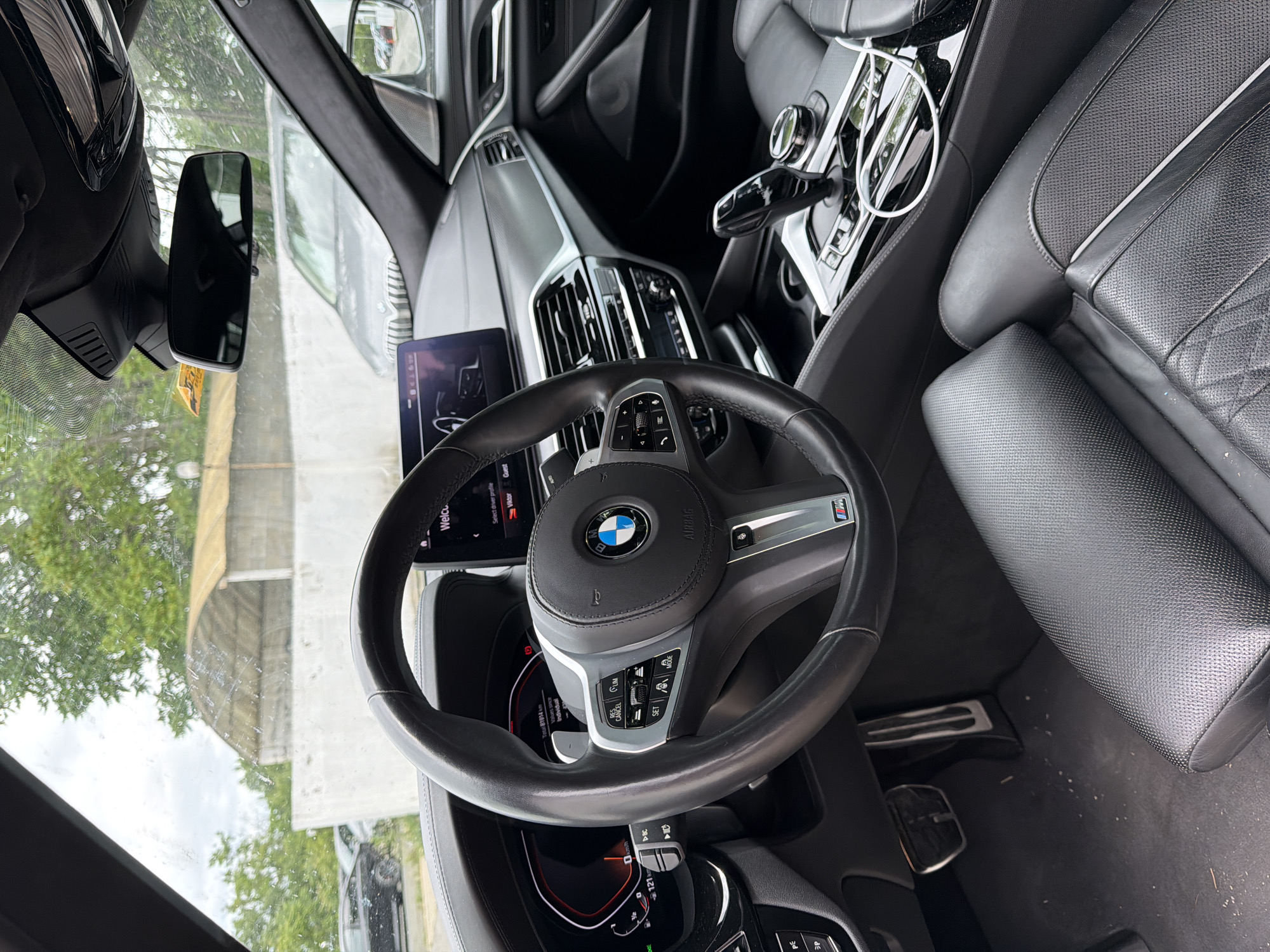 The image shows the interior of a black BMW car. The steering wheel is prominently in the foreground, with the BMW logo visible in the center. The dashboard displays a digital speedometer reading 121 km/h, and a digital screen shows "Welcome" and options to "Select driver profile." The gear shifter and center console are also visible, along with black leather seats. Outside the car, through the windshield and side windows, trees and a cloudy sky are visible. A white building with a curved roof and what appears to be a fence or wall are also present. The car is parked on a paved surface, possibly a parking lot or street. The context of Burgas, Bulgaria is not evident from the visual elements of the image itself.