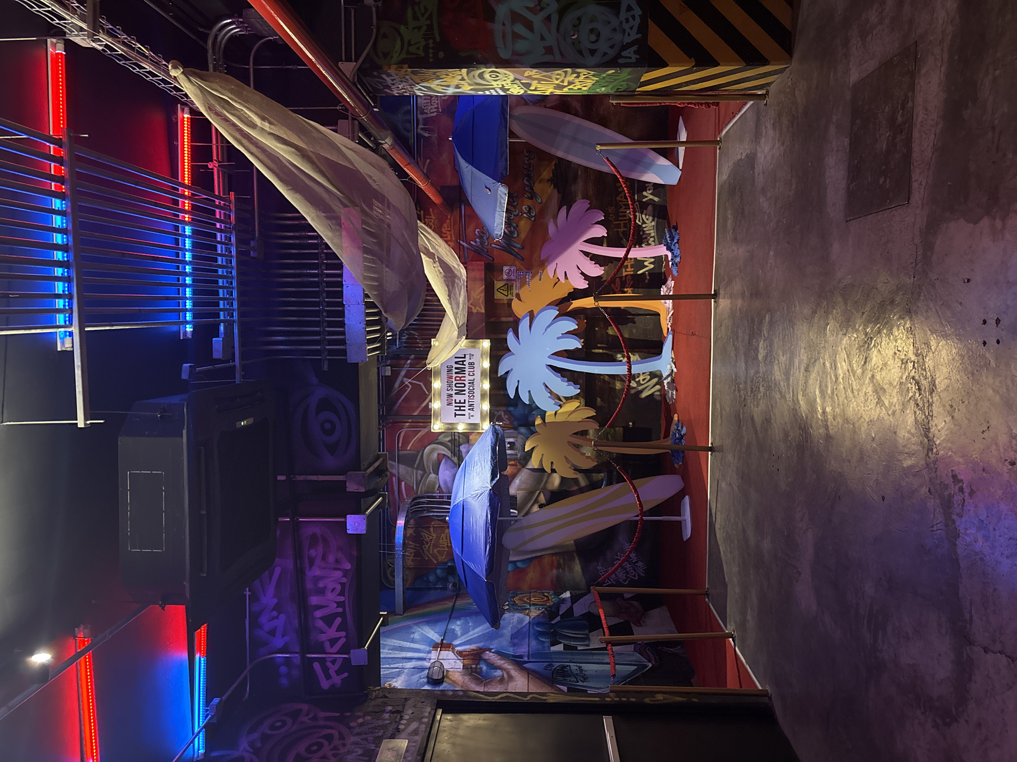Here is a description of the image:
The image presents an indoor scene, possibly a bar or club, with a vibrant and eclectic aesthetic. The space is demarcated by red railings, guiding the viewer's eye towards a wall adorned with graffiti and colorful decorations. Palm tree cutouts in shades of pink, yellow, and blue add a tropical vibe, complemented by surfboards and umbrellas suspended in the air.
The decor is clearly designed to evoke a lively and playful atmosphere. The presence of a sign reading "THE NORMAL ANTISOCIAL CLUB" provides context, suggesting the venue is designed for nightlife and social gatherings. The location, based on your information, would be in San Bernardino Tlaxcalancingo, Mexico.
On the left side of the image, there is a dark area that includes lighting elements with red and blue colors. There is also a black speaker that is near the top of the area. The graffiti on the wall and the use of unconventional decorations such as the umbrellas and surfboards contribute to an urban and creative atmosphere. The floors appear to be concrete and show signs of wear, perhaps adding to a sense of authenticity.