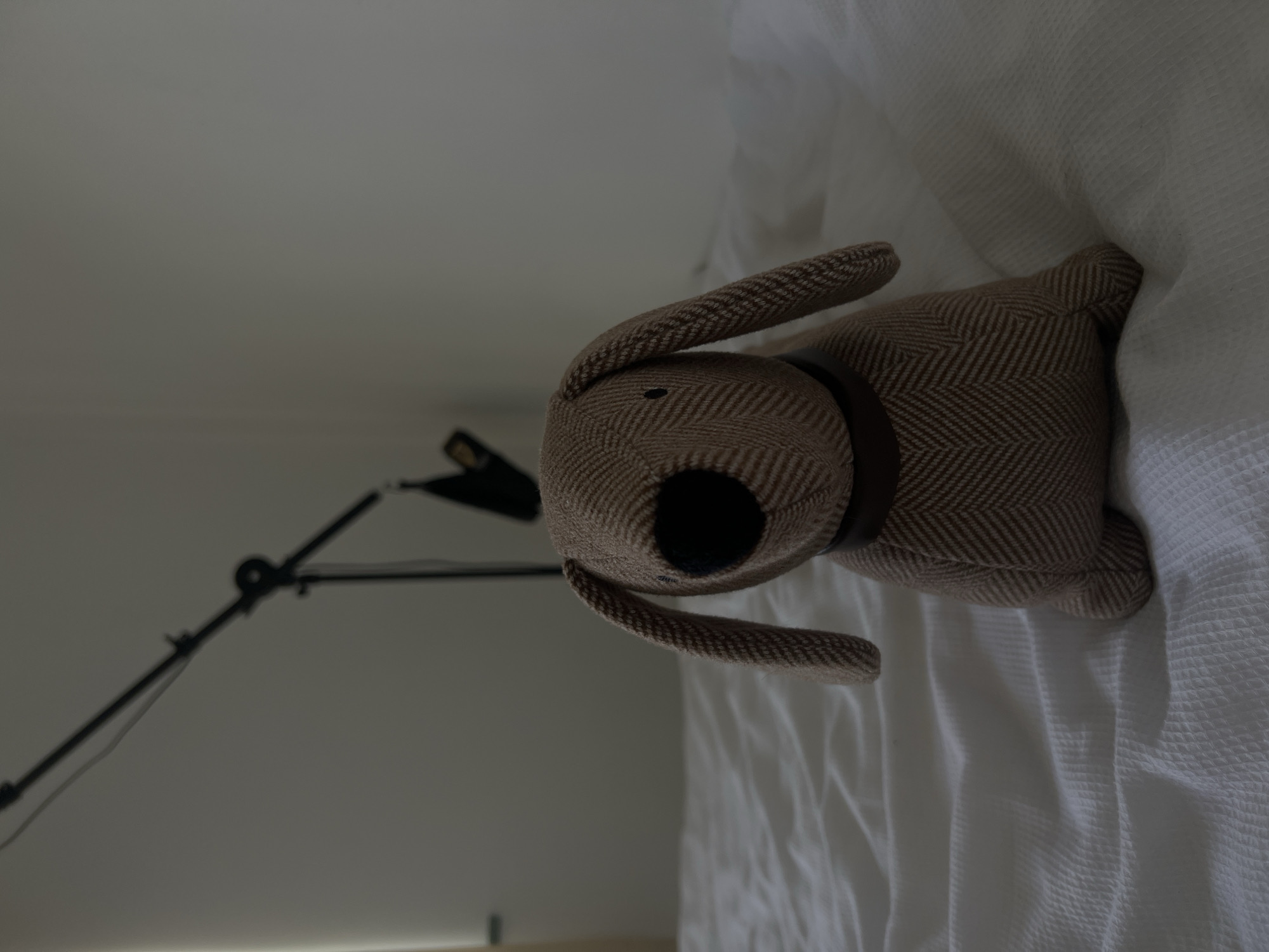 A stuffed dog toy is positioned centrally on a white, textured bedspread. The toy is brown with a herringbone pattern and has long floppy ears, a black nose, and small eyes. It is wearing a dark brown collar. Behind the toy, a black metal stand with an adjustable arm is visible, extending diagonally upwards and to the left, with a black object attached to its end. The background is a plain, light-colored wall. The lighting is dim, casting soft shadows. The scene appears to be indoors, in a bedroom. There is no indication of Balbriggan, Ireland, in the image.