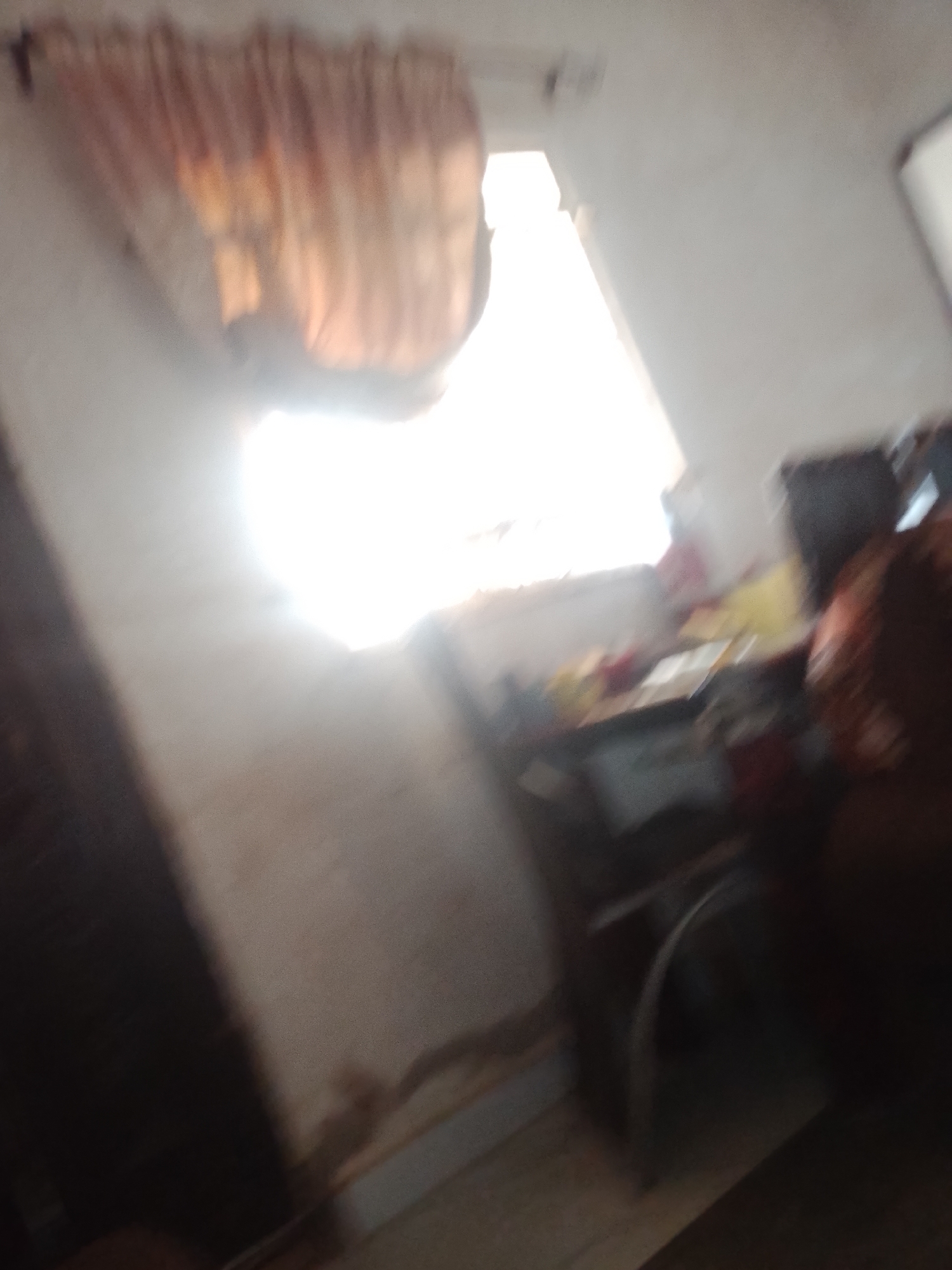 The image shows an interior scene with a window that is brightly lit by sunlight. There are curtains visible above the window, which appear to be made of fabric with a striped pattern. Below the window, there is a desk or table with various items on it, though they are not clearly discernible due to the blurriness of the image. A dark object, possibly furniture or a person, is visible to the right of the desk. The walls appear to be light-colored and show some texture or discoloration. The overall impression is that of a somewhat cluttered indoor space with natural light coming from the window. The scene is located in the city of Jalingo, Nigeria.