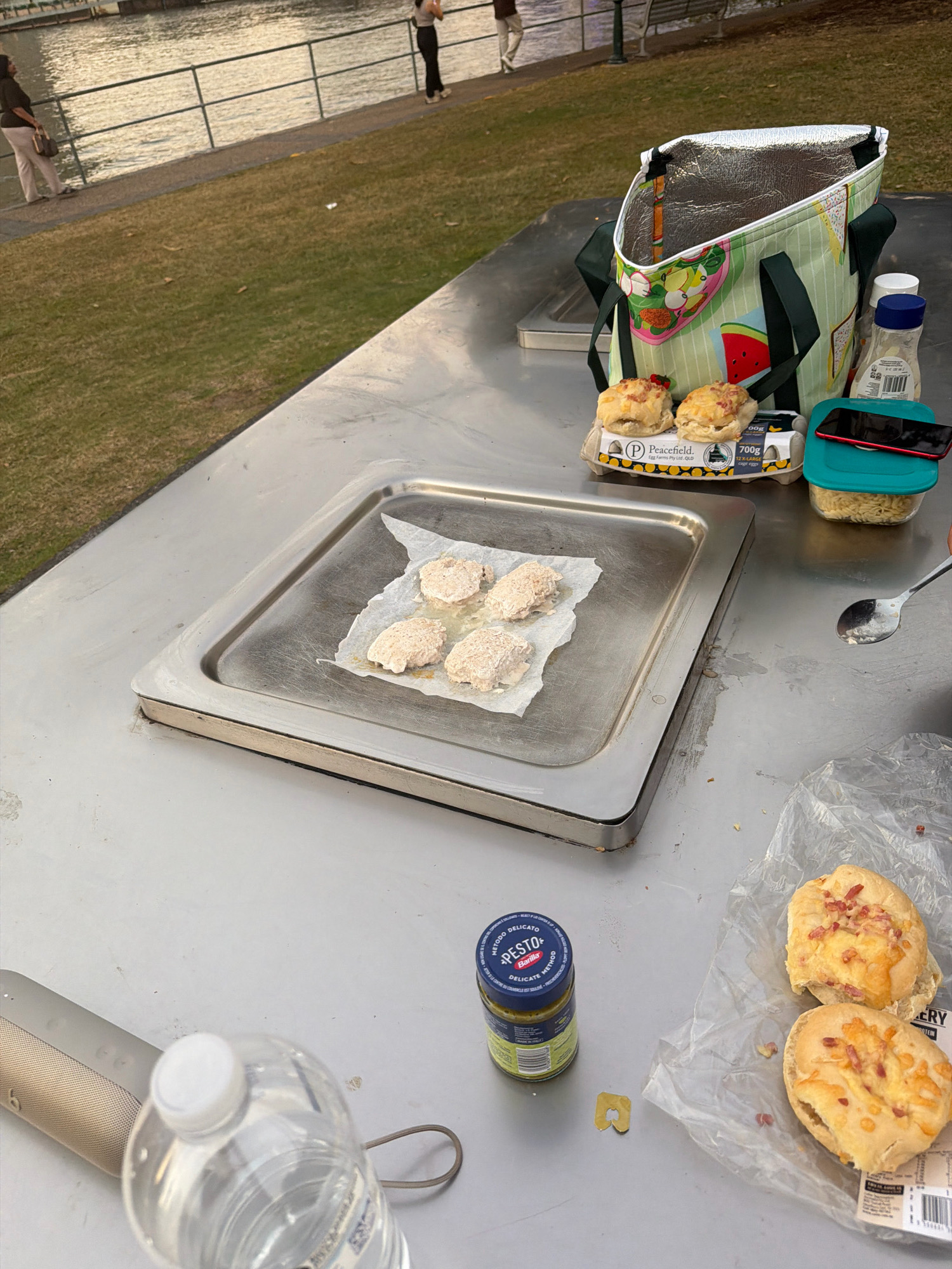 An outdoor dining scene is depicted at Kangaroo Point, Australia, with the Brisbane River visible in the background. The foreground shows a stainless steel table laden with food items and accessories for a picnic. In the center, a tray holds what appear to be four breaded patties, possibly chicken tenders, arranged on parchment paper, ready for cooking. To the right, a cooler bag with a fruit pattern is open, revealing its insulated interior. Beside it, a carton of eggs from "Peacefield" is partially visible, with two small bread rolls topped with cheese and bacon pieces placed on top. A jar of "Pesto" by Barilla stands next to the egg carton. A smartphone with a red case rests on a teal plastic container. A water bottle with a white cap is also present, along with a portable speaker. In the background, people are seen strolling along a path beside the river, with a railing separating them from the water. The setting appears to be a park or public recreational area, suggesting a leisurely outdoor meal.